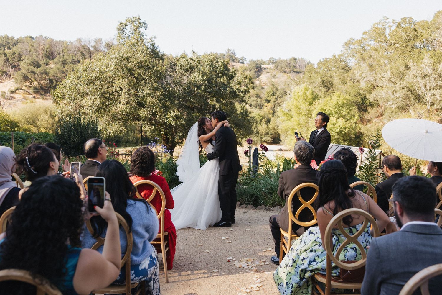Wedding guests watching ceremony in a rustic outdoor setting with wooden chairs and natural surroundings.