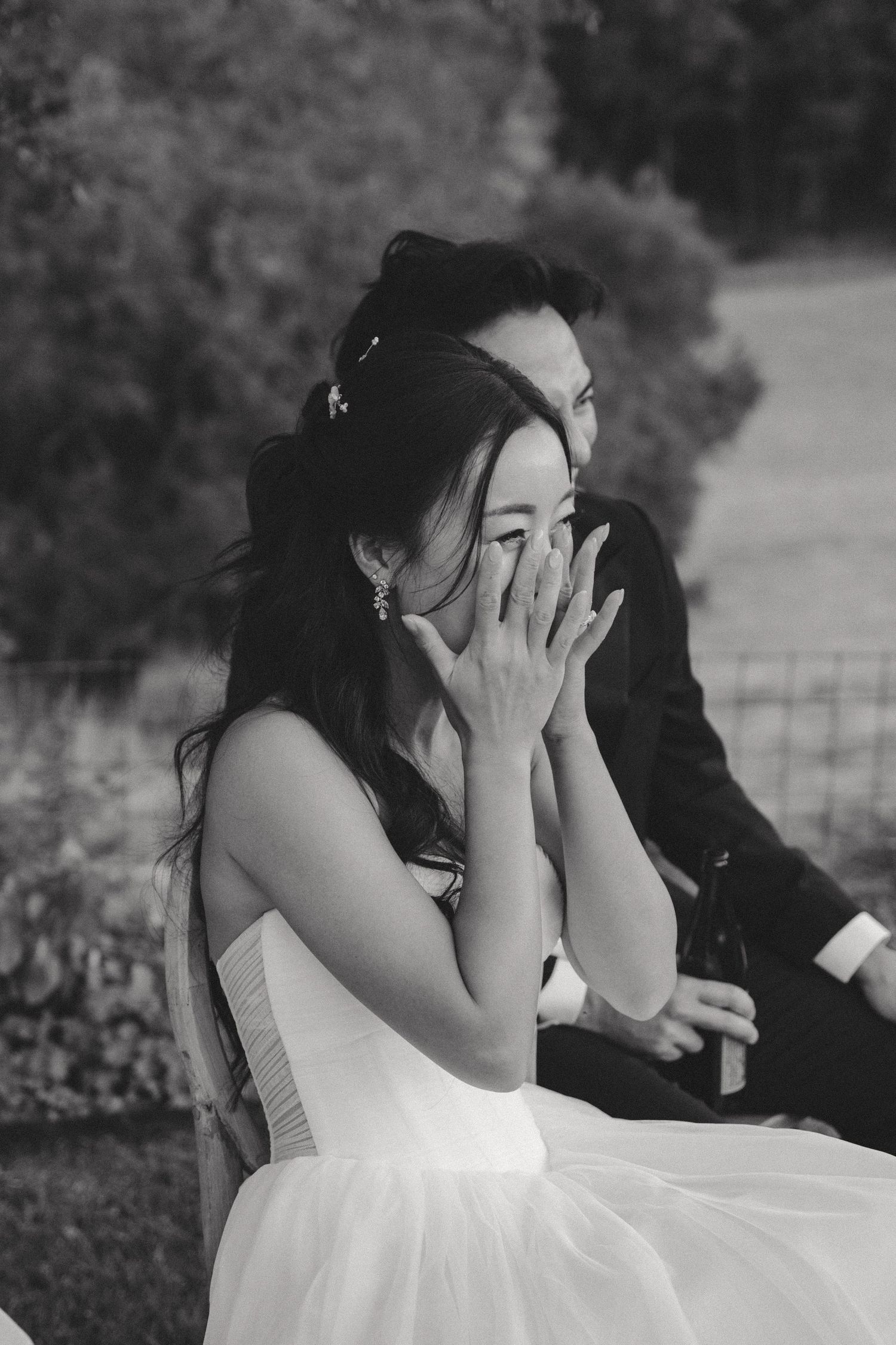 A person in a white dress covers their face with their hands in an emotional moment outdoors.