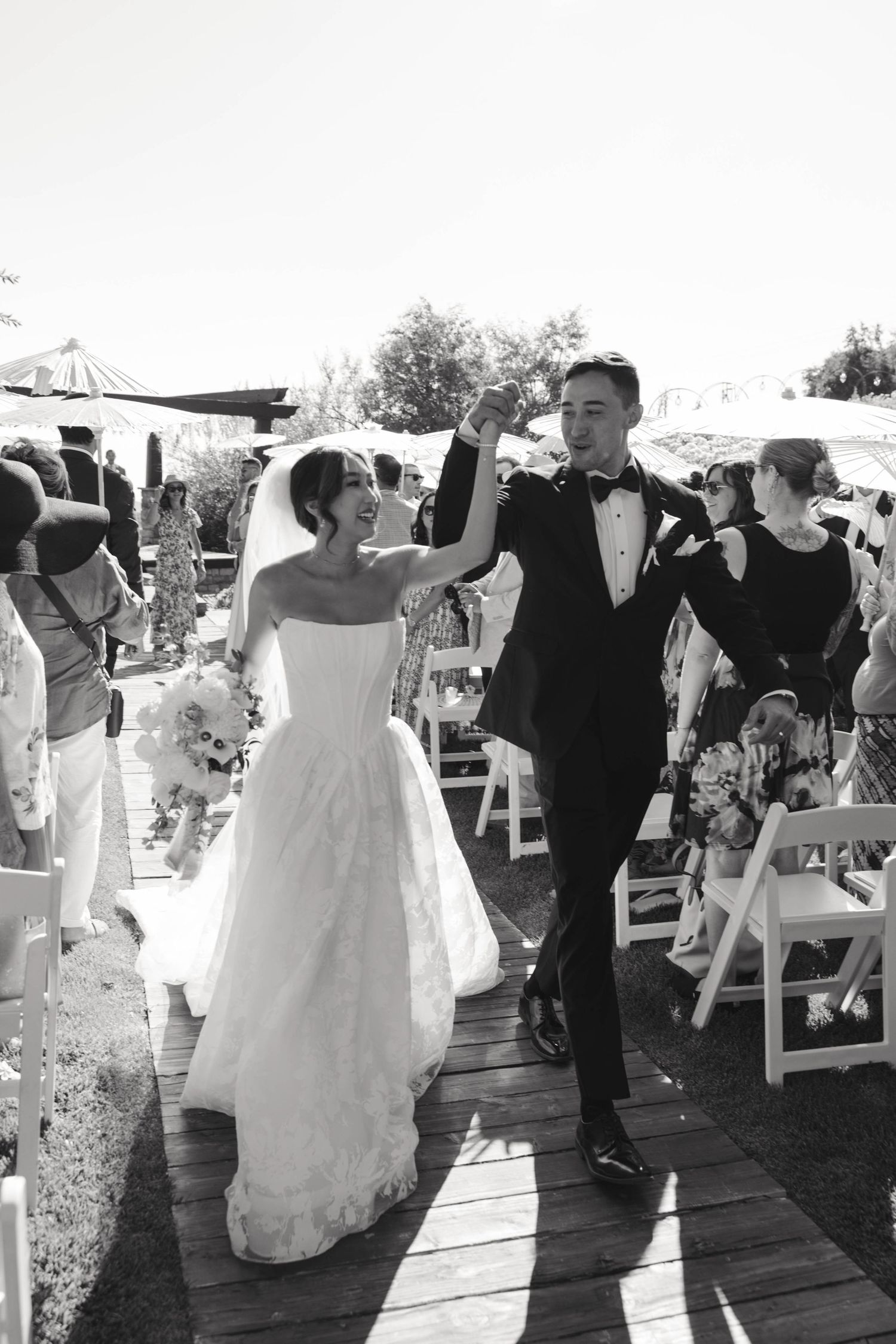 A wedding ceremony recessional with a couple joyfully walking down the aisle among white chairs at an outdoor venue.