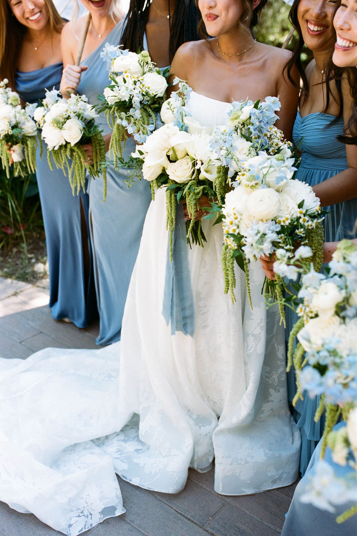 Wedding party holding white and blue floral bouquets while wearing dusty blue bridesmaid dresses.