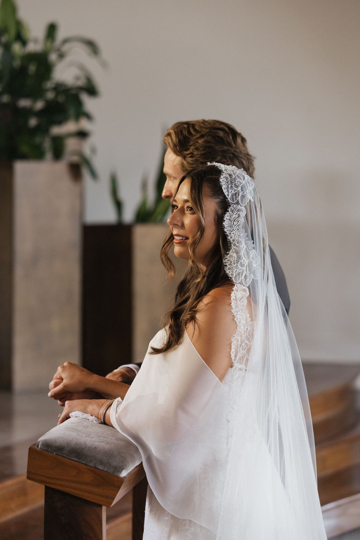 Bride in white gown and lace veil kneeling at church pew, smiling softly during wedding ceremony.