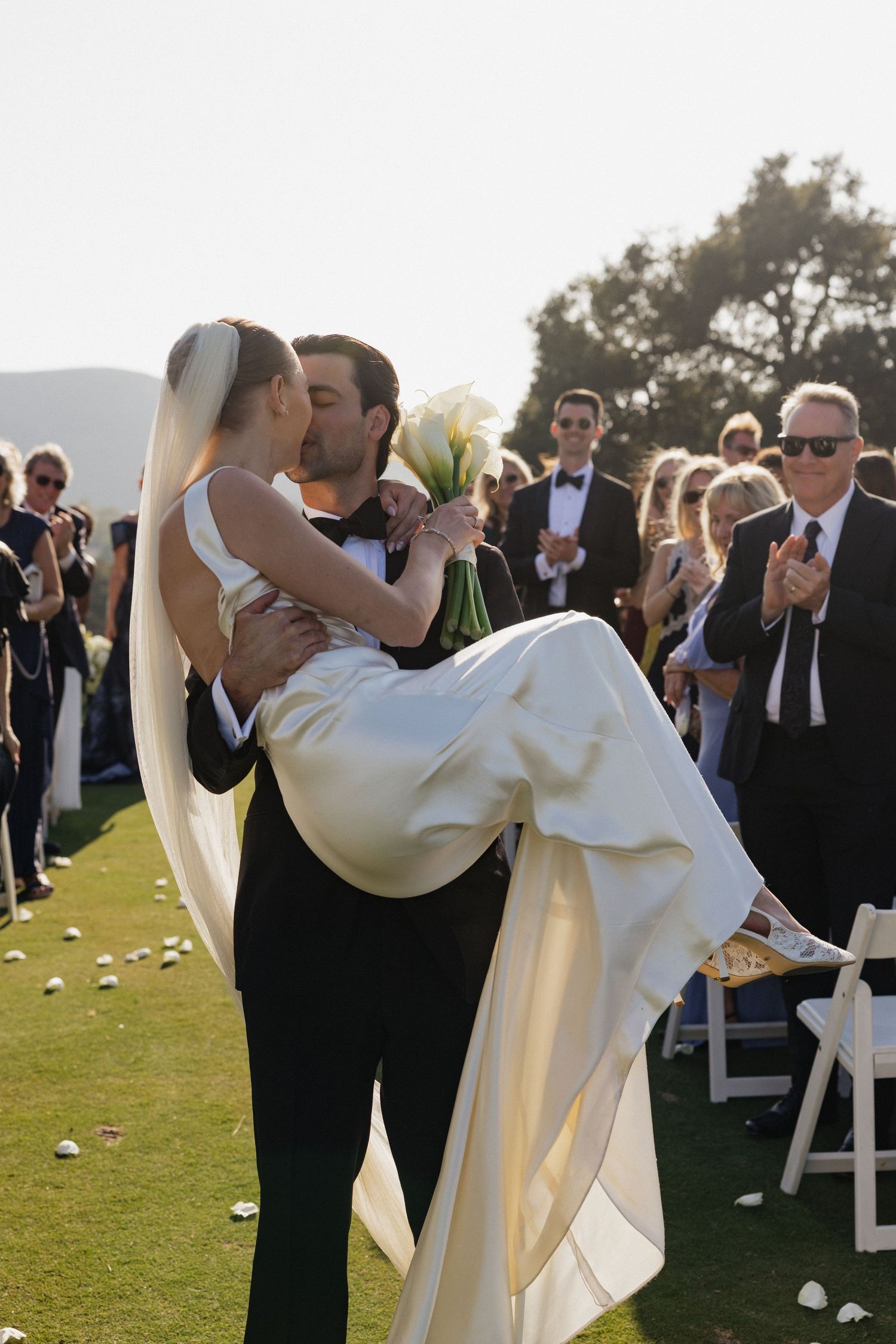 Groom lifts bride in white gown and veil, sharing a kiss at outdoor wedding ceremony with guests applauding.