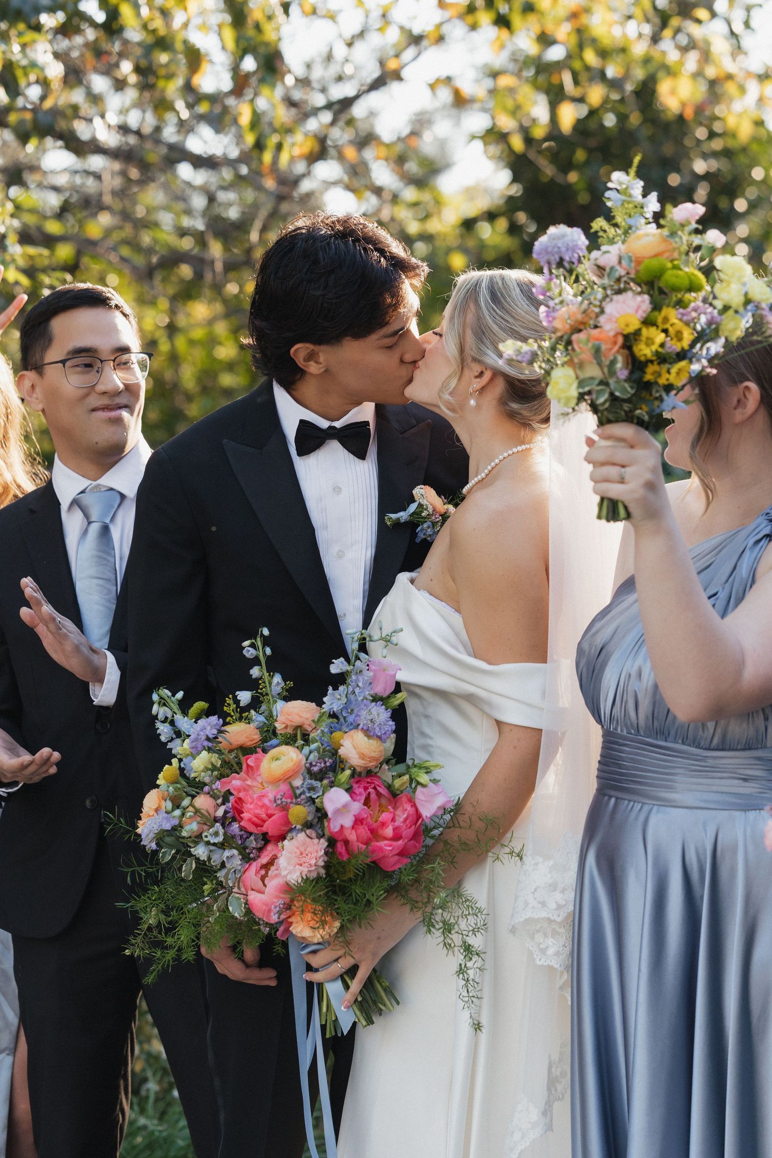 Wedding party members in formal attire with light blue bridesmaid dresses and colorful floral bouquets stand together outdoors.