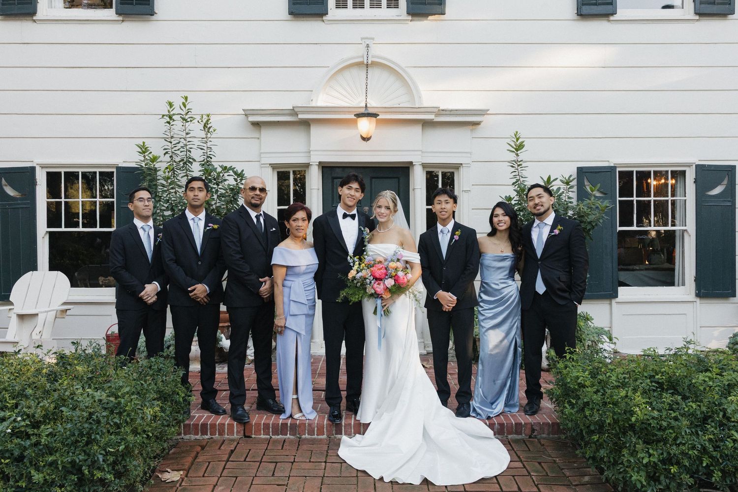 Wedding party poses in front of white colonial-style building with black shutters and string lights overhead.