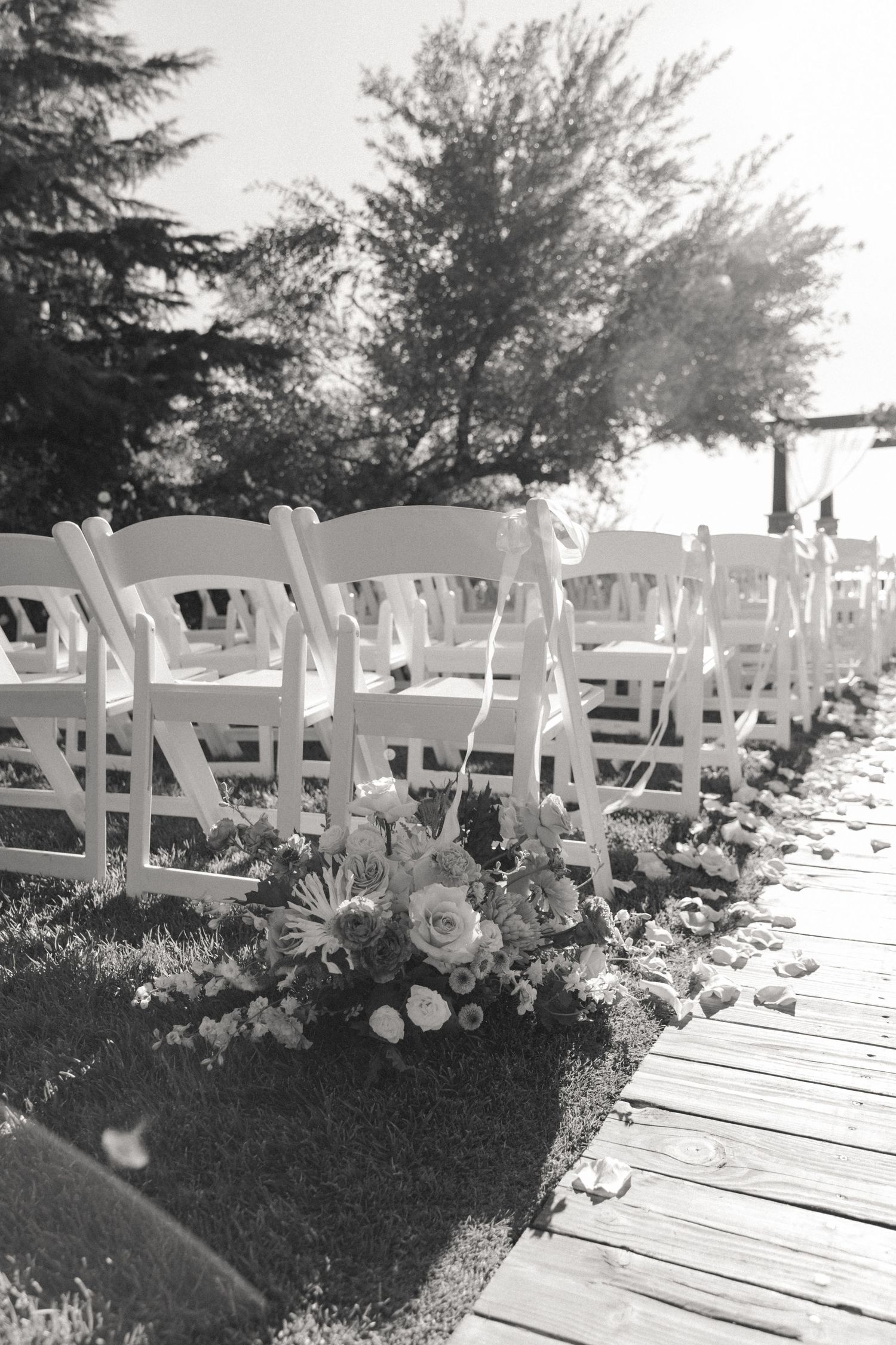 White wedding ceremony chairs line a wooden walkway with floral arrangements and trees in the background.