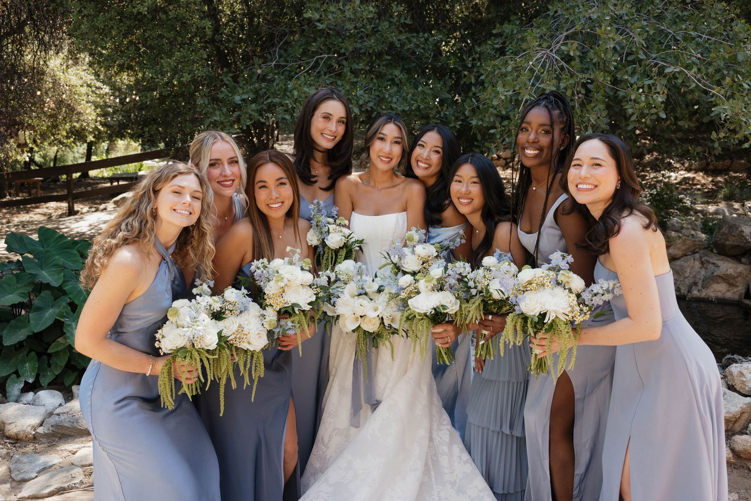 Wedding party in light blue bridesmaid dresses poses with bouquets outdoors on a sunny day.