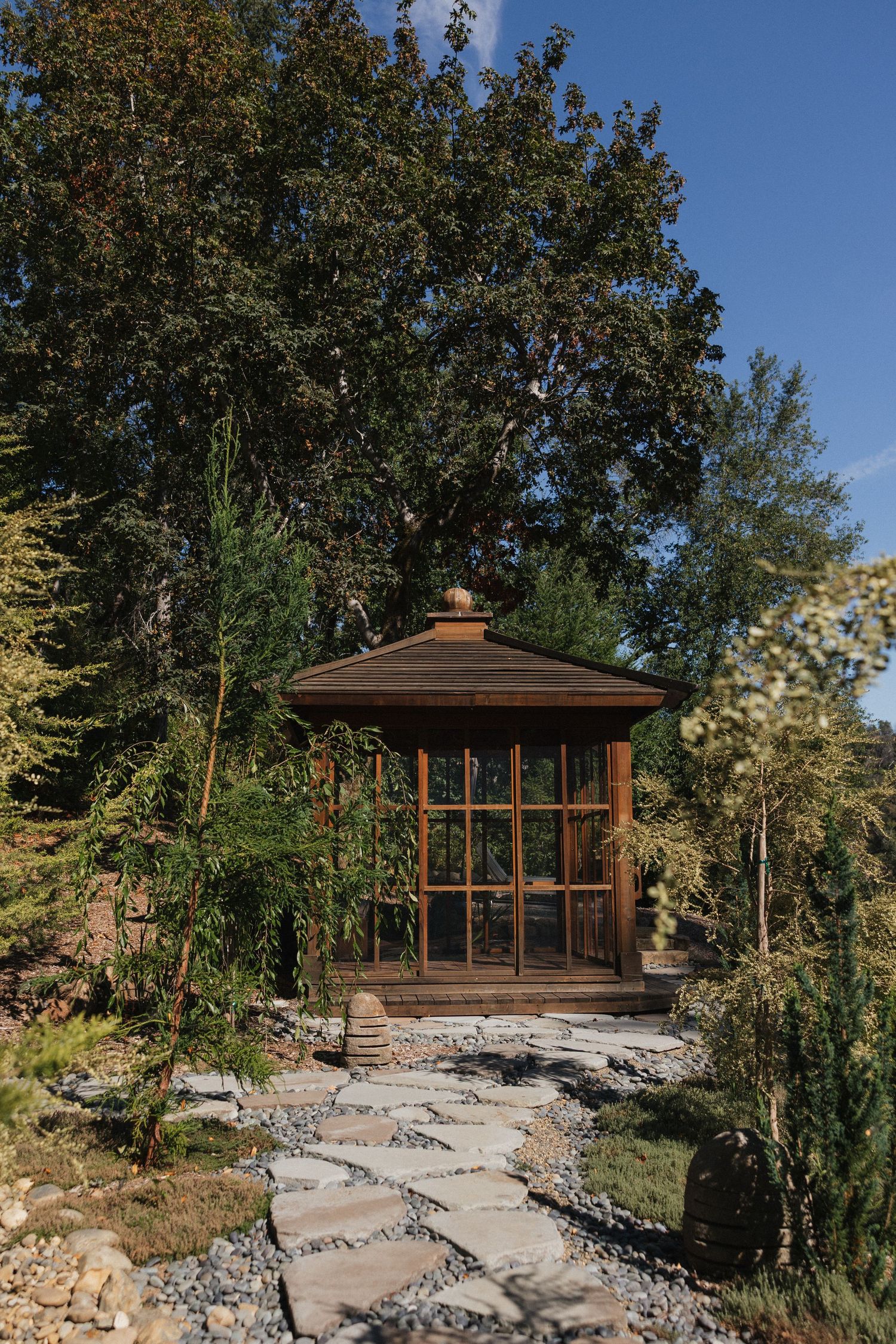 Wooden gazebo with glass windows sits atop stone landscaping surrounded by trees in a tranquil outdoor setting.