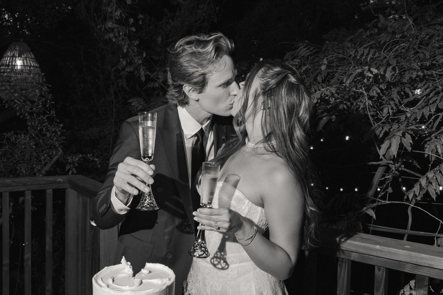 Black and white photo of a couple kissing while holding champagne glasses at an outdoor wedding reception.