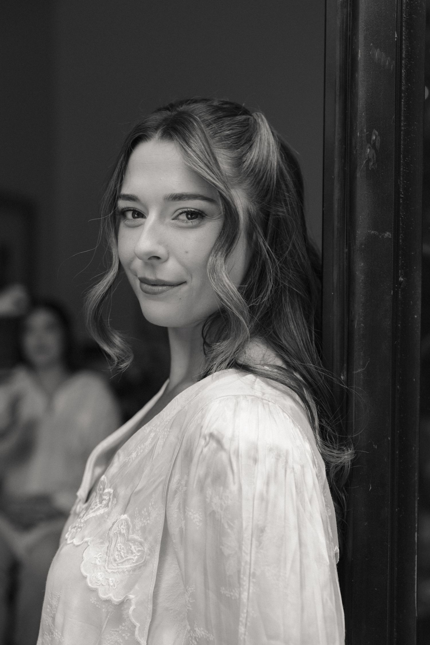 Black and white portrait of a young woman in a lace blouse leaning against a dark doorframe.