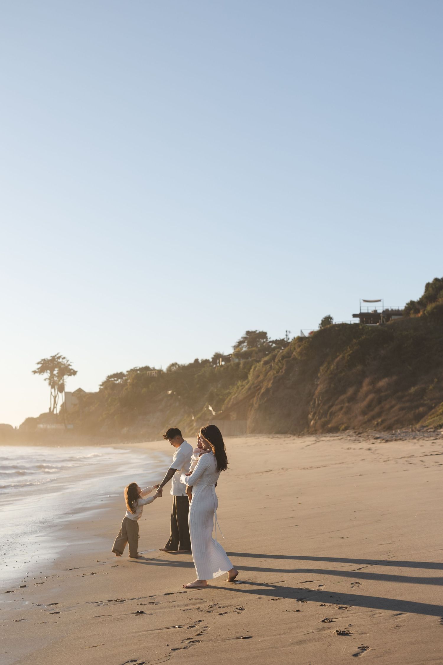 A couple in white outfits walks with a small child on a scenic beach at sunset with cliffs in the background.