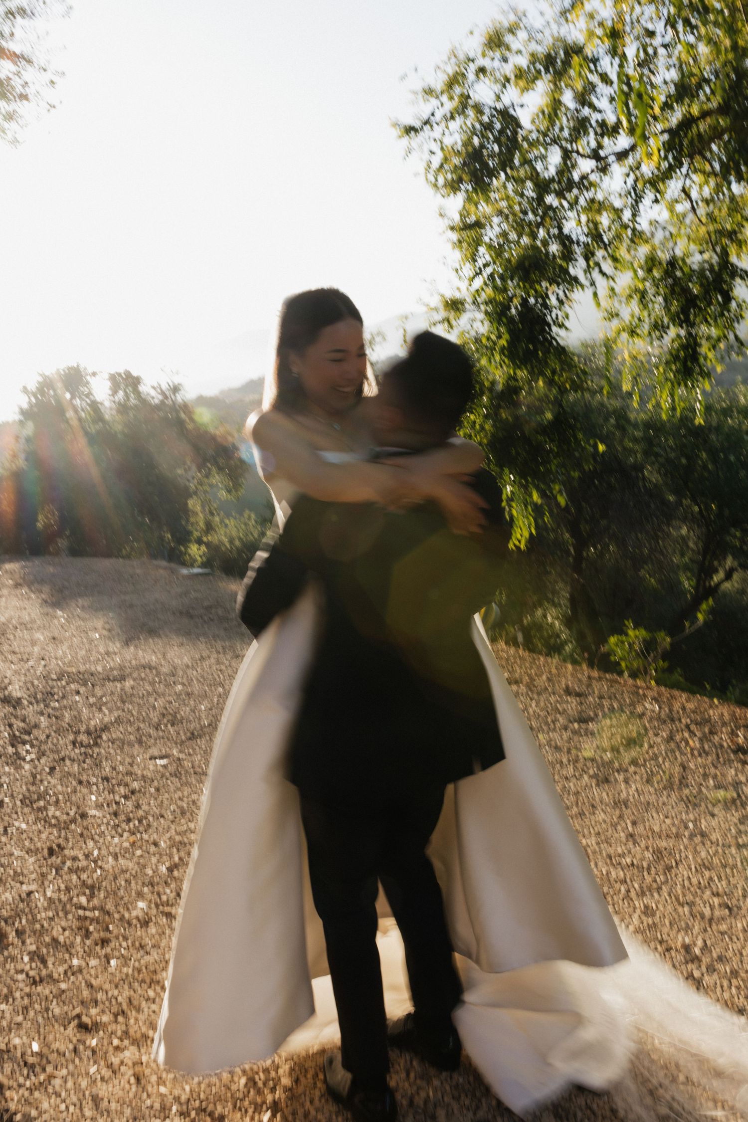 Groom lifting bride in white gown outdoors, sun-drenched wedding photo with lush greenery backdrop.