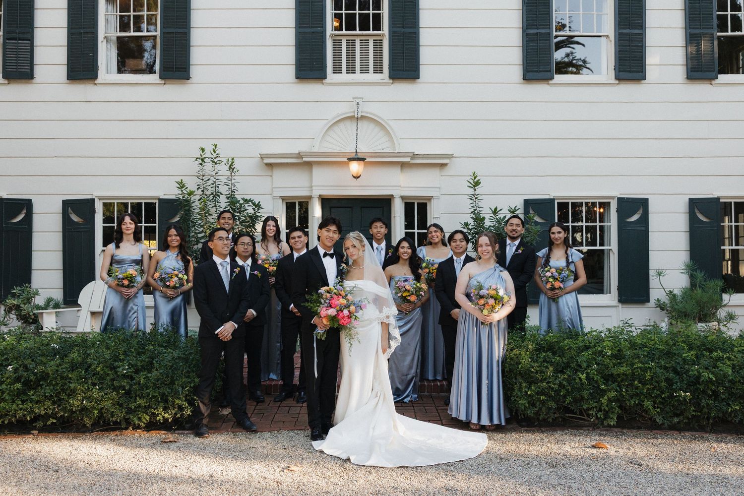 Wedding party members stand together in formal attire against classic white building exterior.