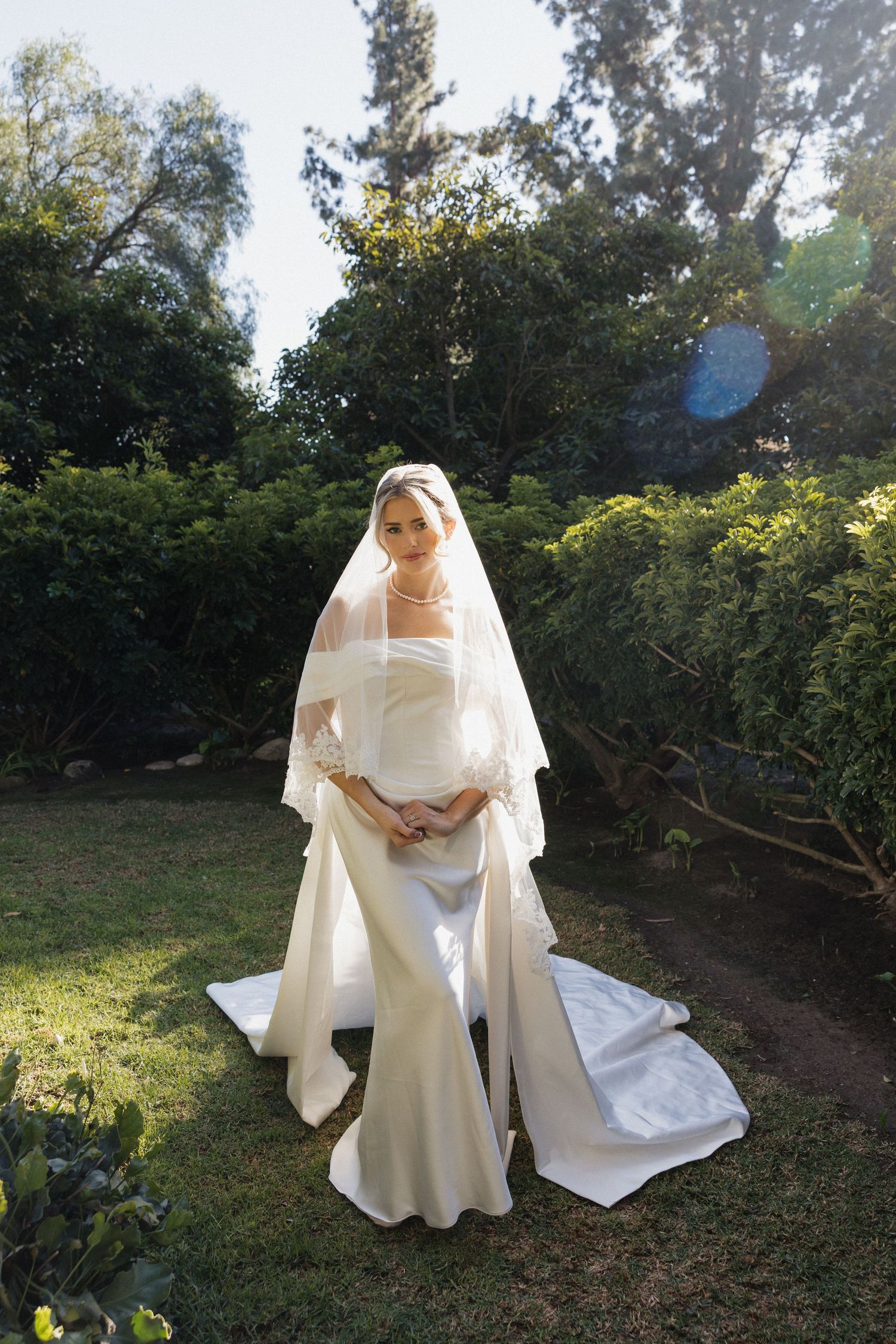 Elegant bridal portrait session in a garden setting with flowing white dress and veil at sunset.