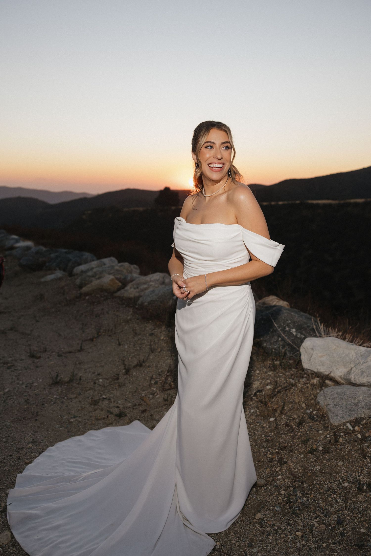 A bride in an elegant off-shoulder gown poses against a mountain sunset backdrop.