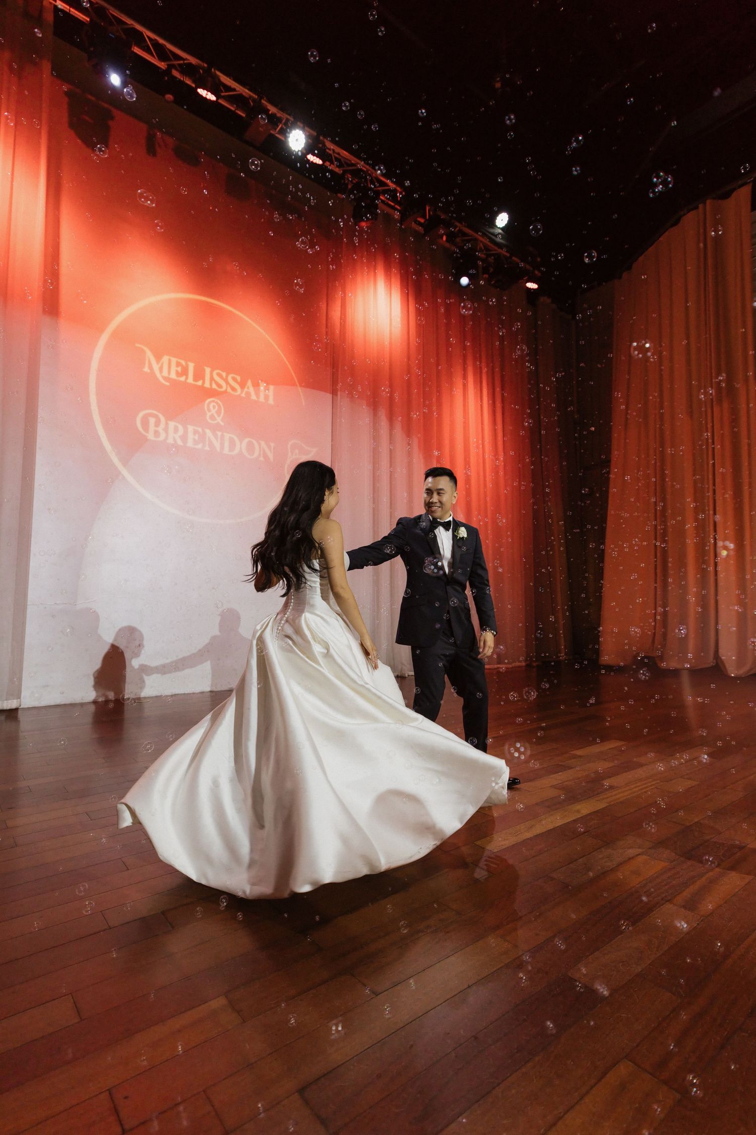 Bride and groom share first dance on wooden stage with red curtains and monogram backdrop at elegant wedding reception.