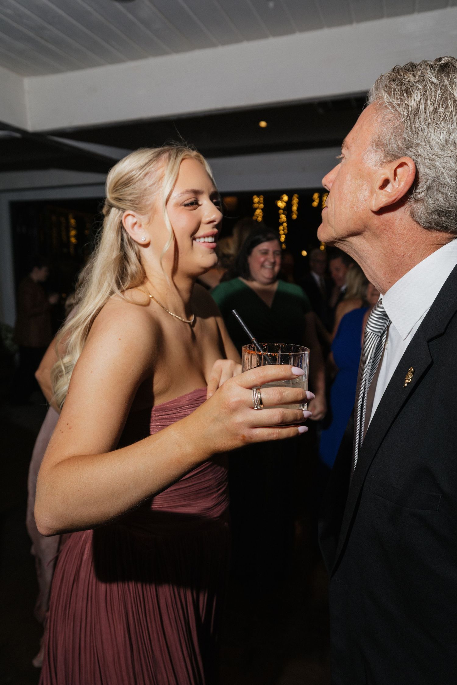 Blonde woman in mauve dress laughing with older man in suit at wedding reception.