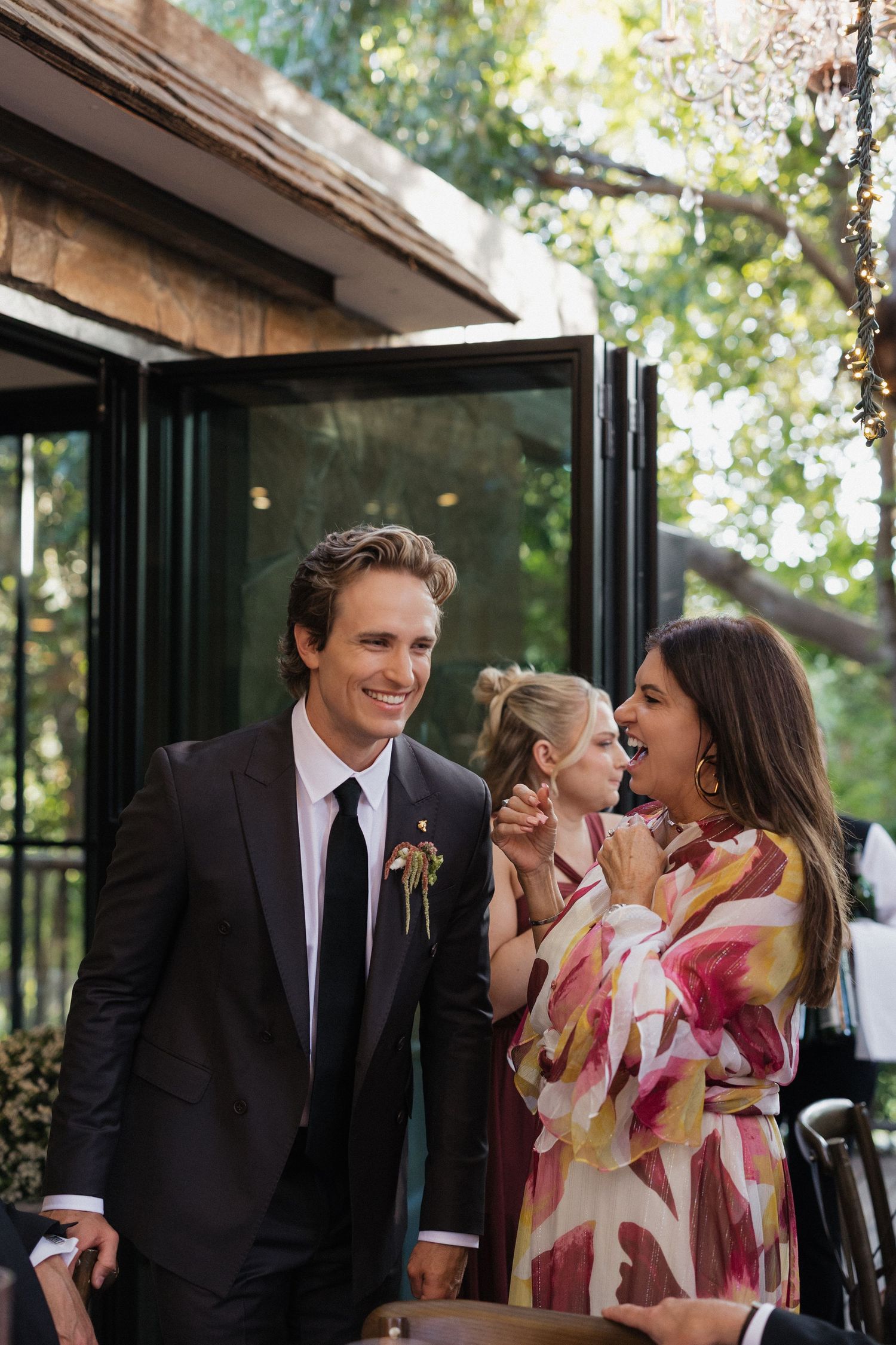 Groom in black suit smiling with guest in floral dress at outdoor wedding reception.