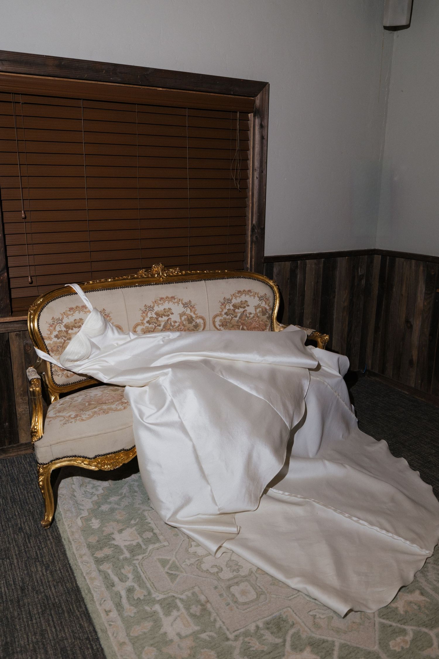 A white wedding dress draped over an ornate gold and cream French Louis XVI style sofa on a floral rug.