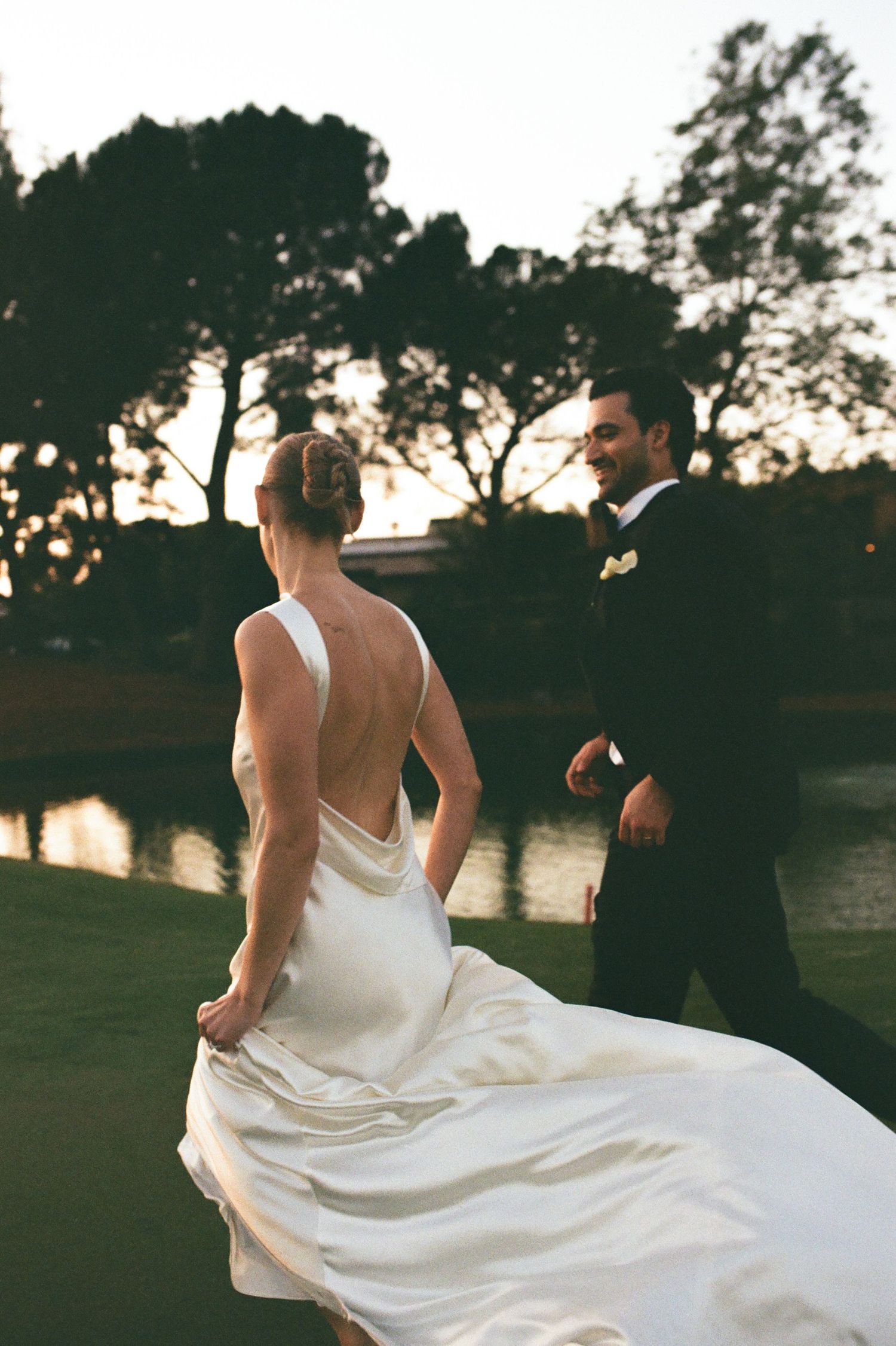 Bride in backless white gown and groom in tuxedo walk together at sunset near a pond.