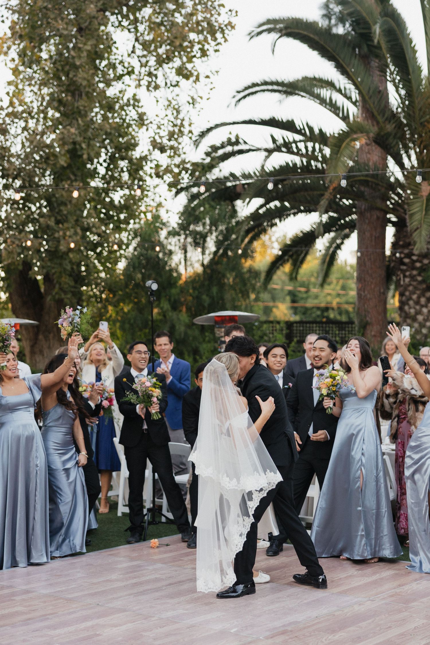 Wedding party in grey and black attire celebrates during an outdoor reception near palm trees.