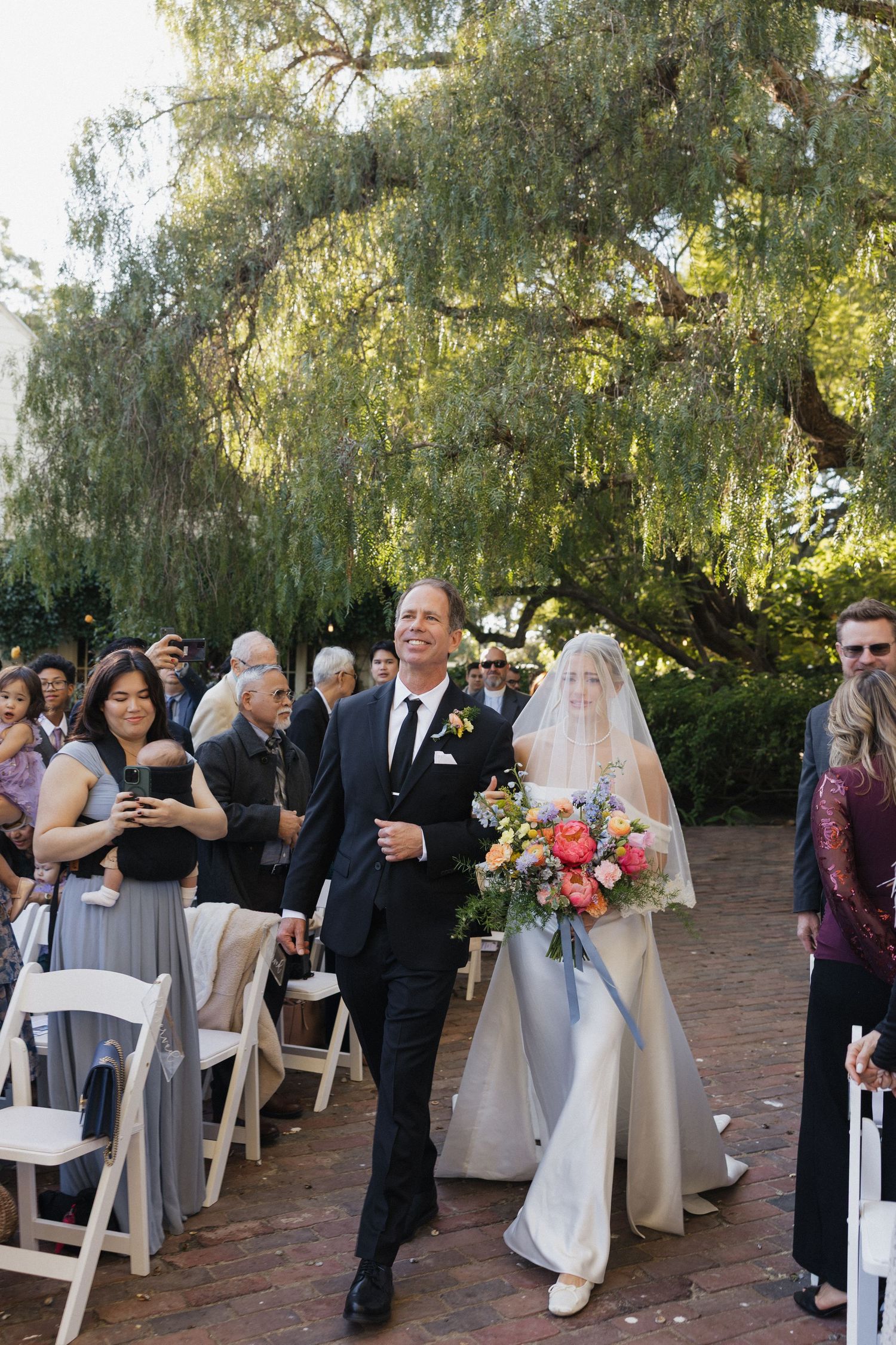 Wedding ceremony guests gather under willow trees with white chairs arranged in rows for an outdoor celebration.