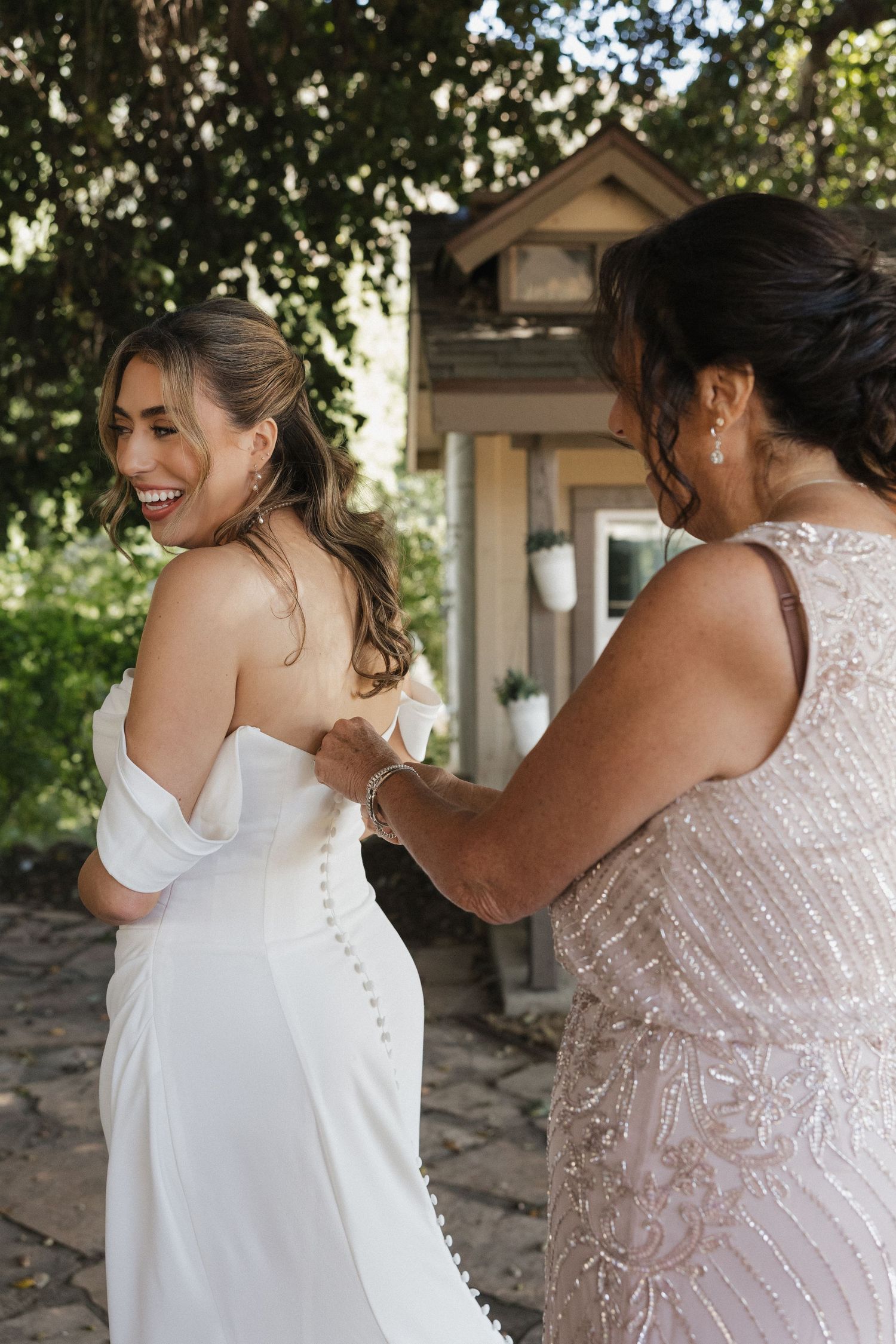 A wedding dress being buttoned up during preparation with delicate lace details in a sunlit outdoor setting.