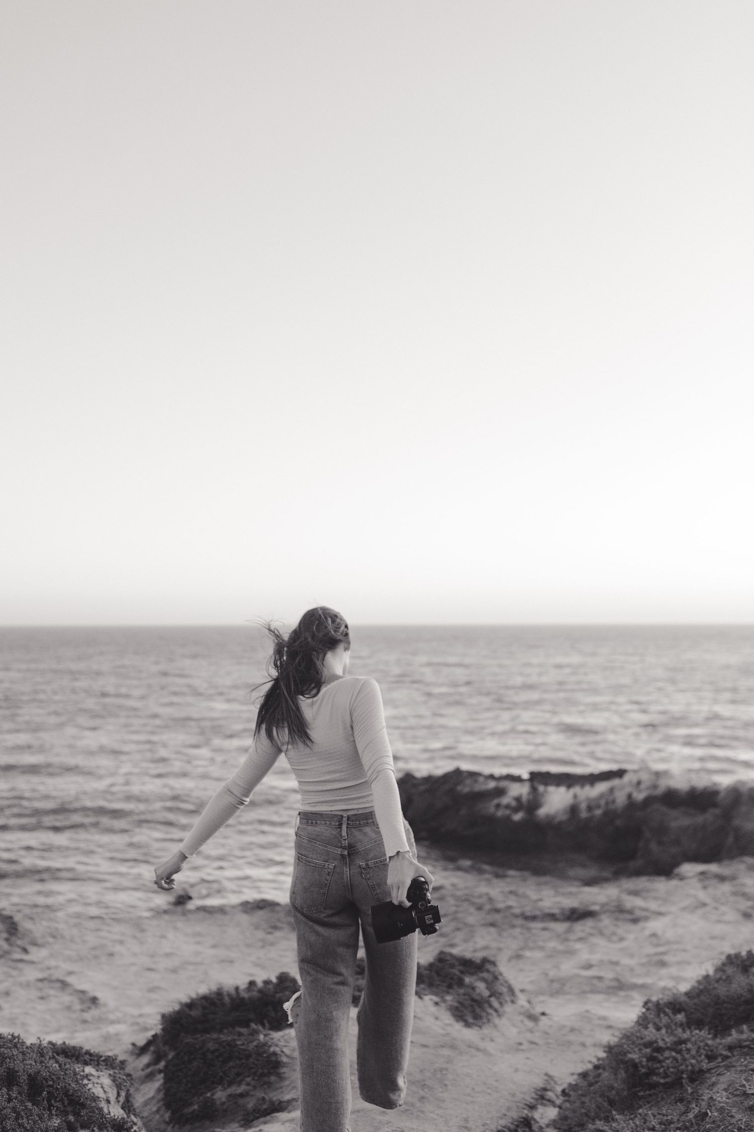 A silhouetted figure stands on a rocky coastline at sunset holding a camera, overlooking the ocean waves.