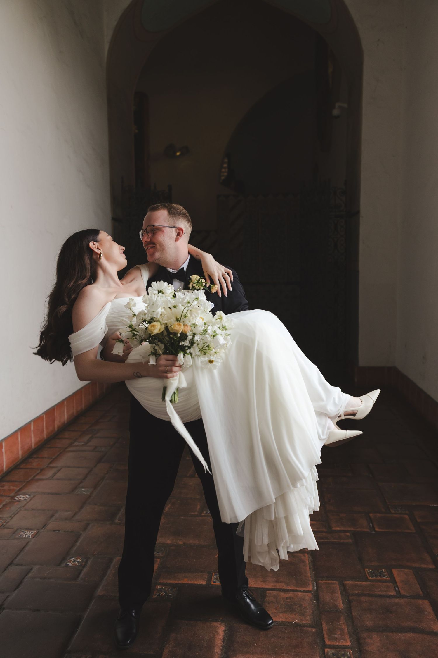 A romantic wedding couple embraces in an arched hallway with terracotta flooring as the groom carries his bride.