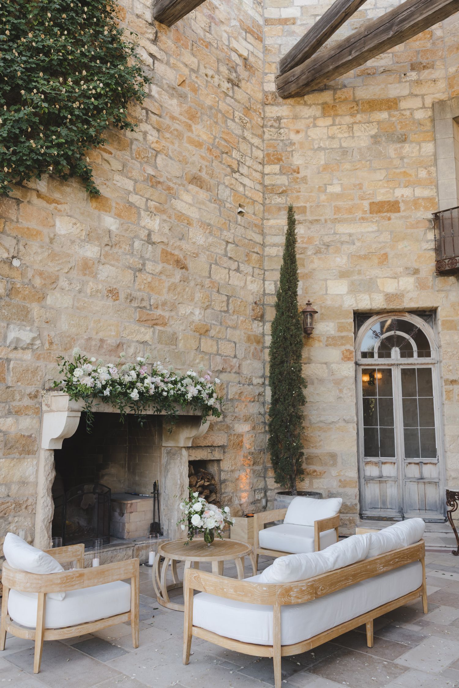 An elegant outdoor seating area with cream cushioned furniture against a stone wall with floral arrangements.