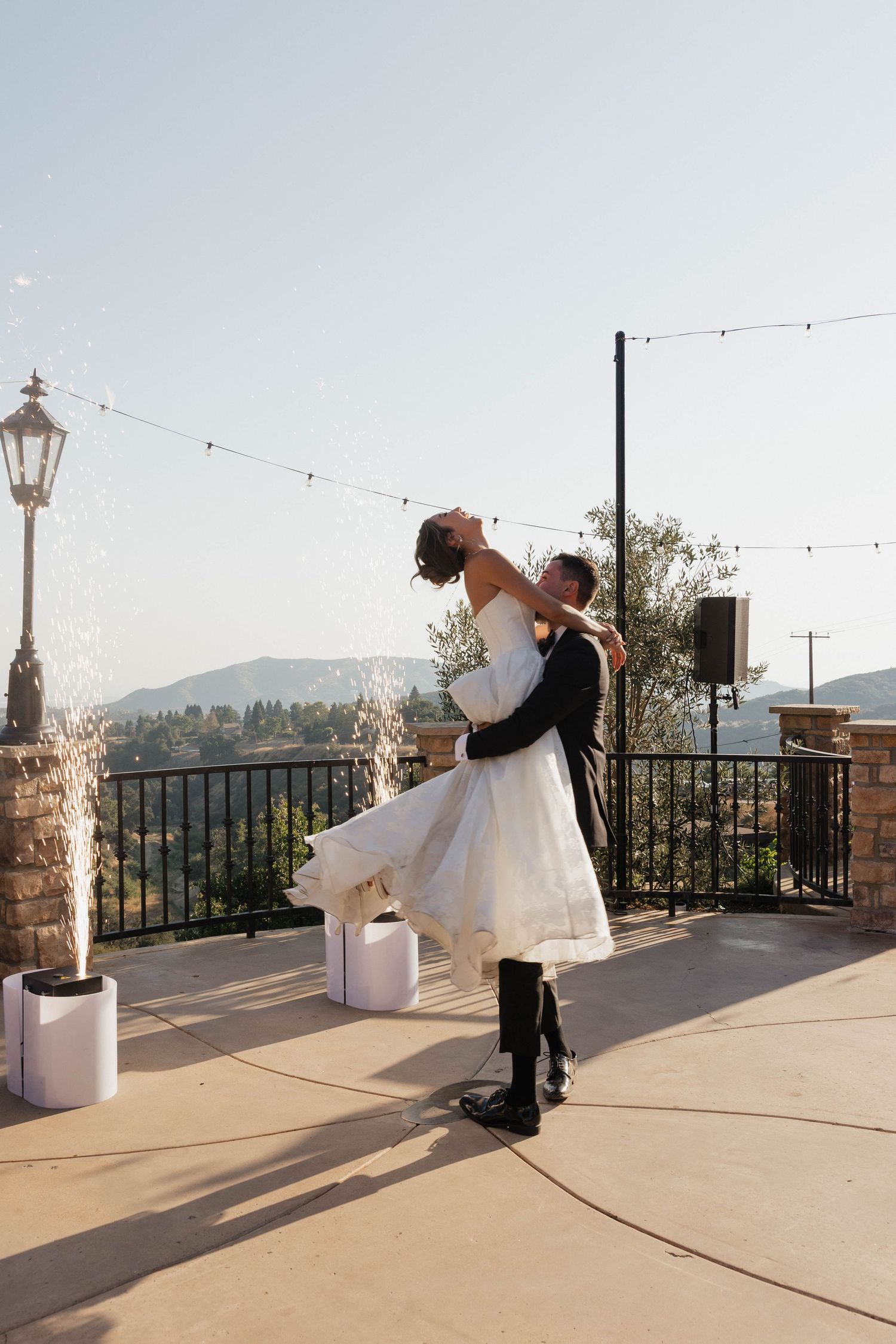 Romantic lift pose between newlyweds on scenic mountain terrace with string lights.