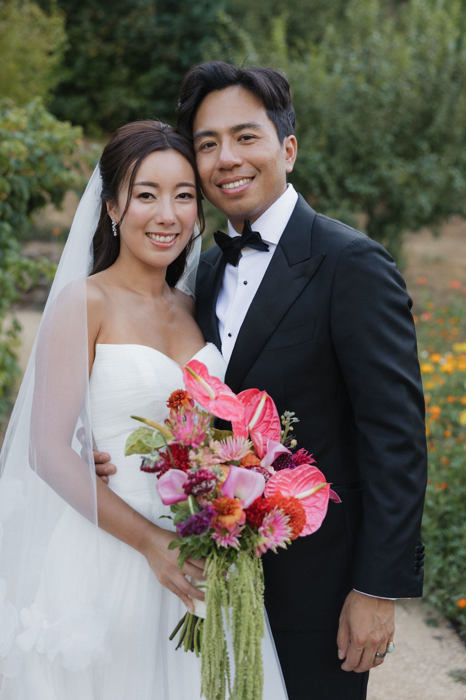 A wedding couple poses outdoors holding a vibrant pink and purple floral bouquet.