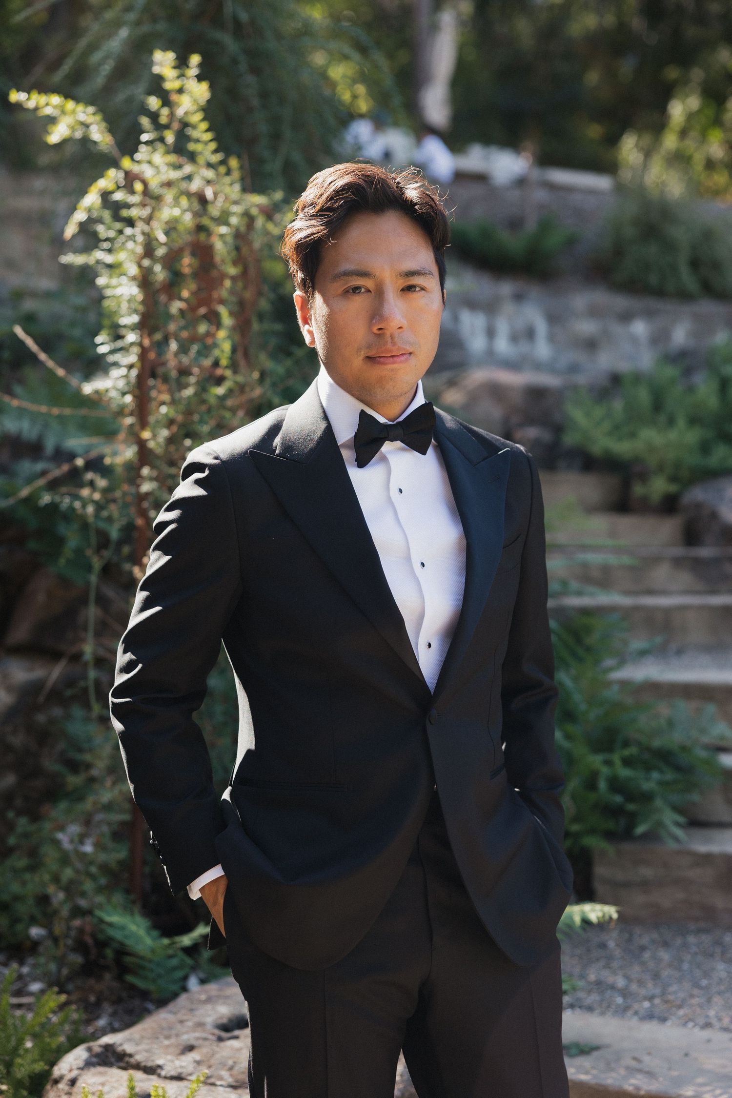 Person in formal black tuxedo and white bowtie poses in a garden setting with natural sunlight.