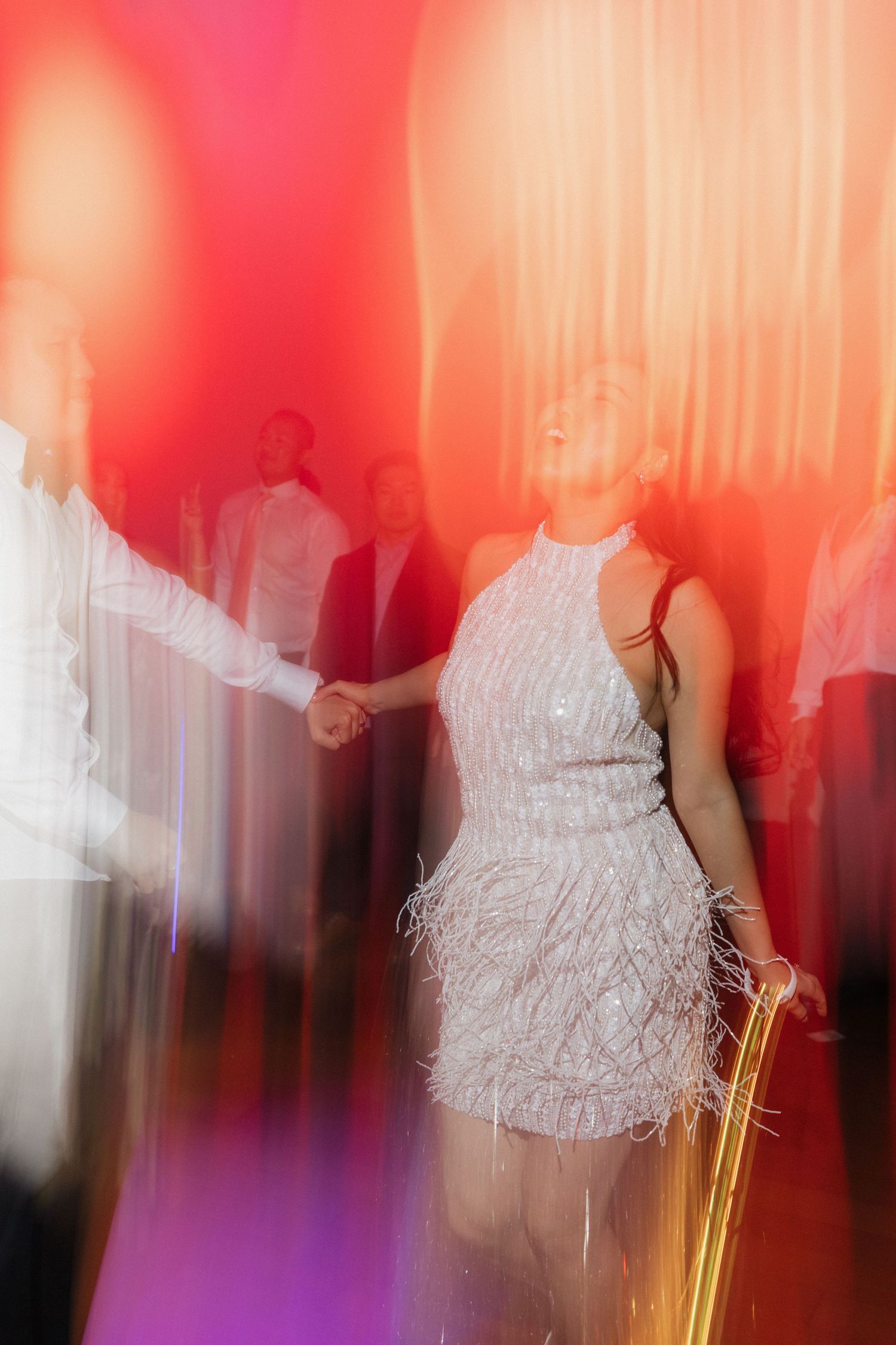 Woman in white feathered mini dress dancing joyfully under colorful red and purple lights at an elegant event.