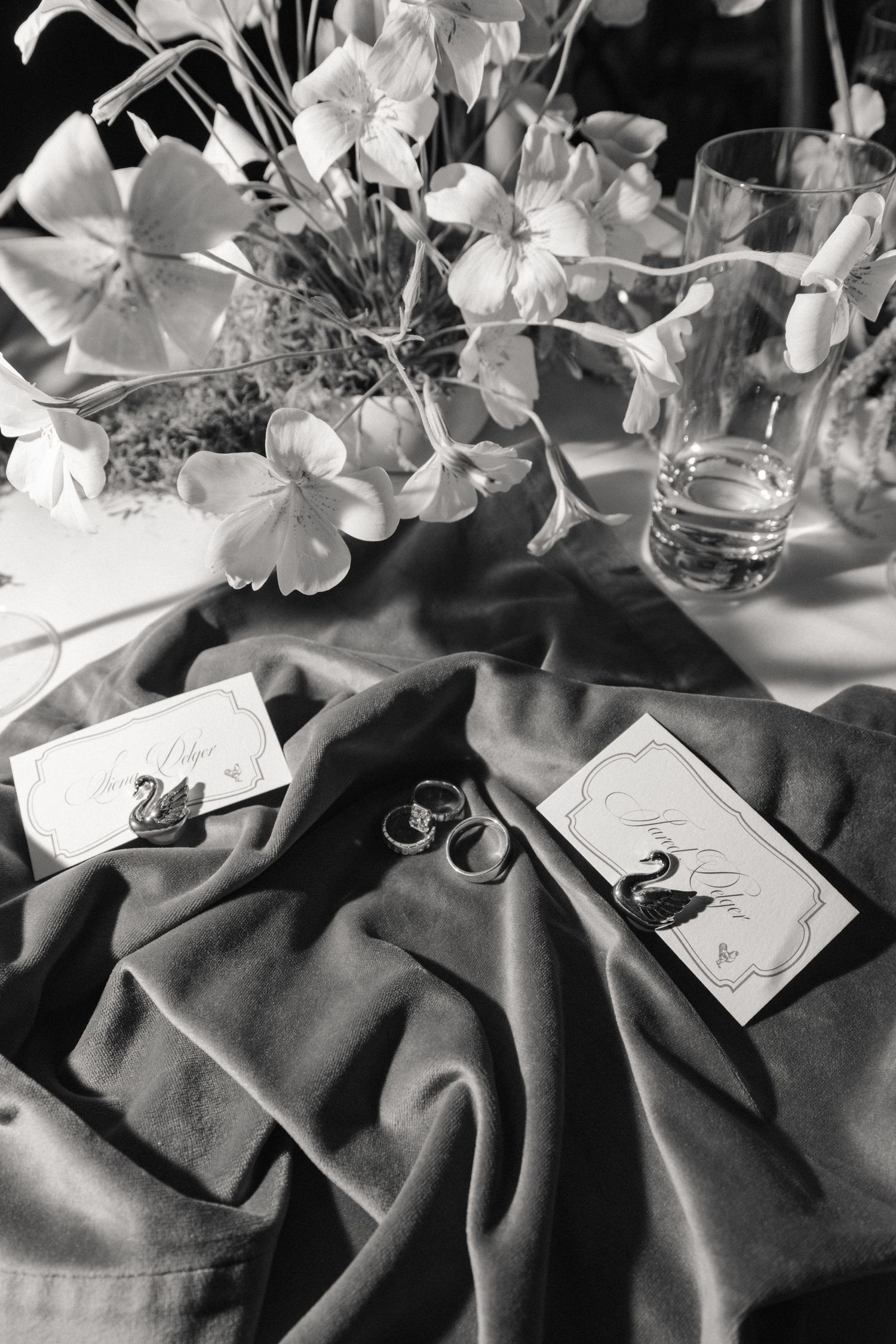 Black and white flat lay of wedding rings and swan place cards on draped fabric with flowers.