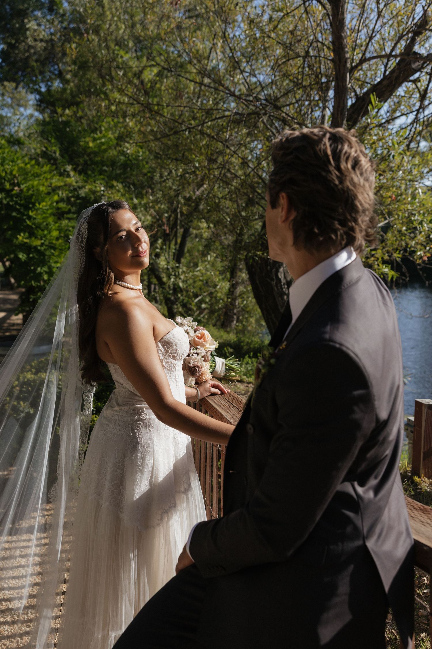 Bride in strapless gown with veil holds bouquet while gazing at groom on wooden bridge outdoors.