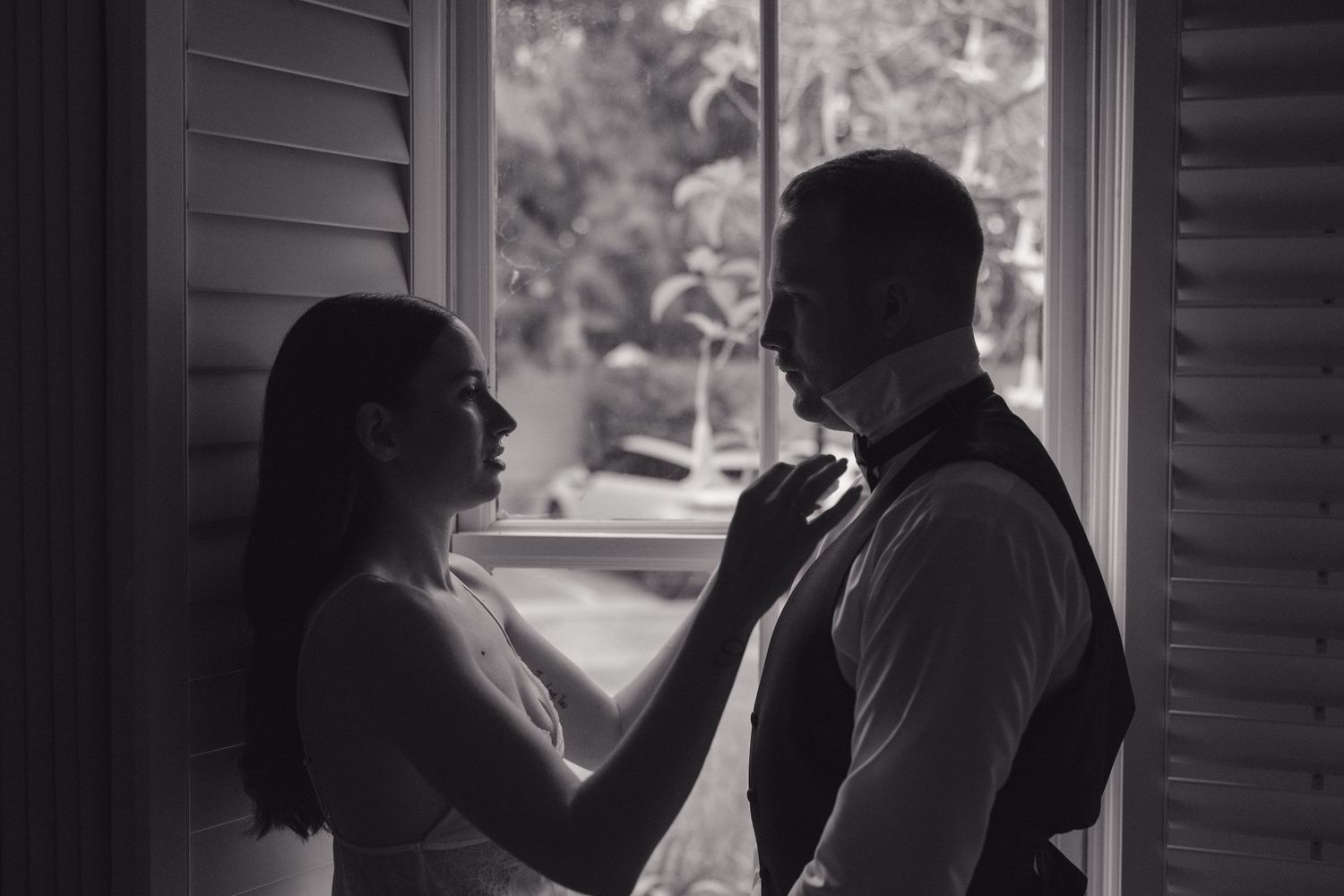 Black and white photo of a couple sharing an intimate moment by a window with natural light streaming in.