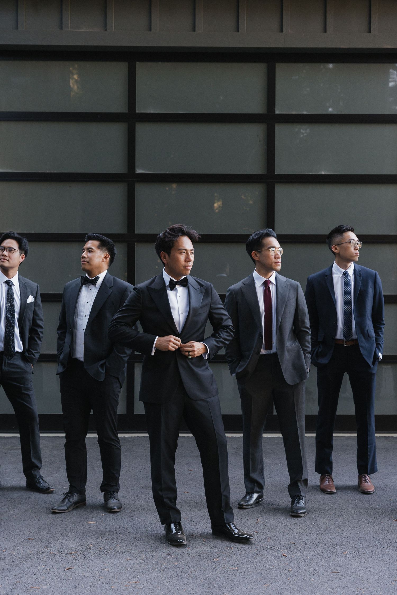 Groomsmen in dark suits pose casually against a modern grey wall.