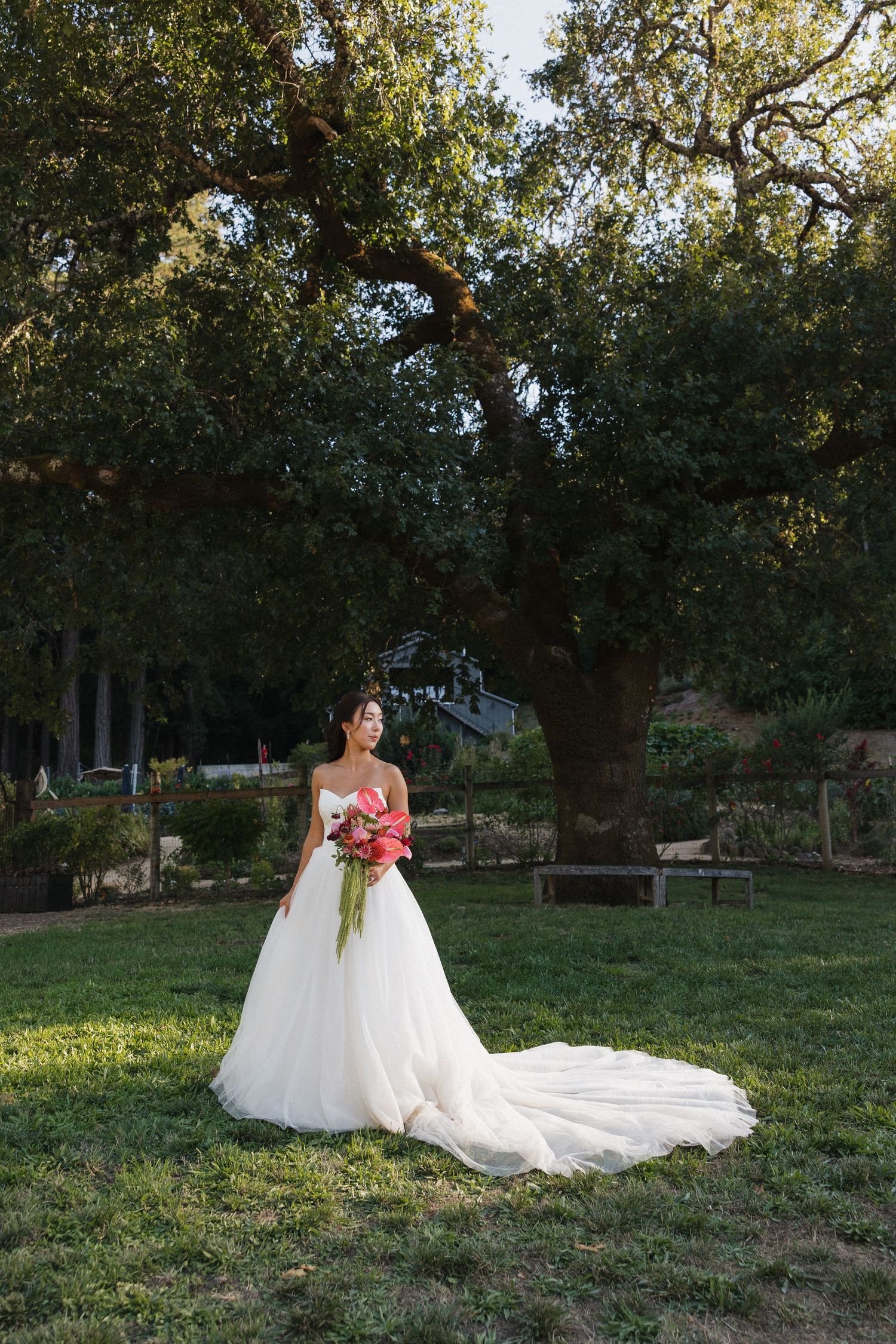 A bride in a flowing white gown holds a bouquet while standing in a garden setting.