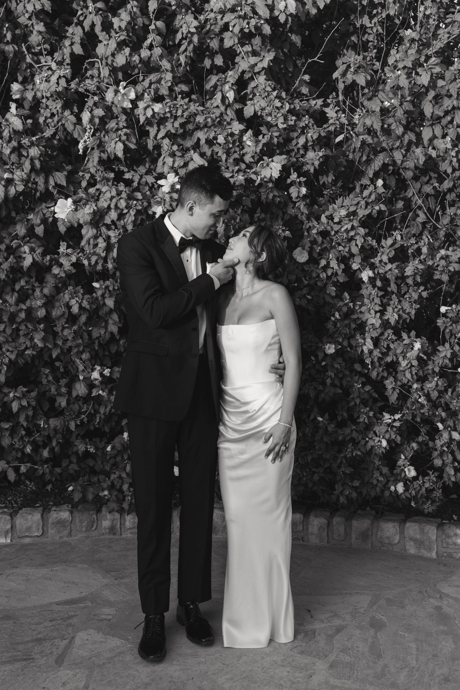 Black and white photo of a couple sharing an intimate moment against a leafy backdrop.