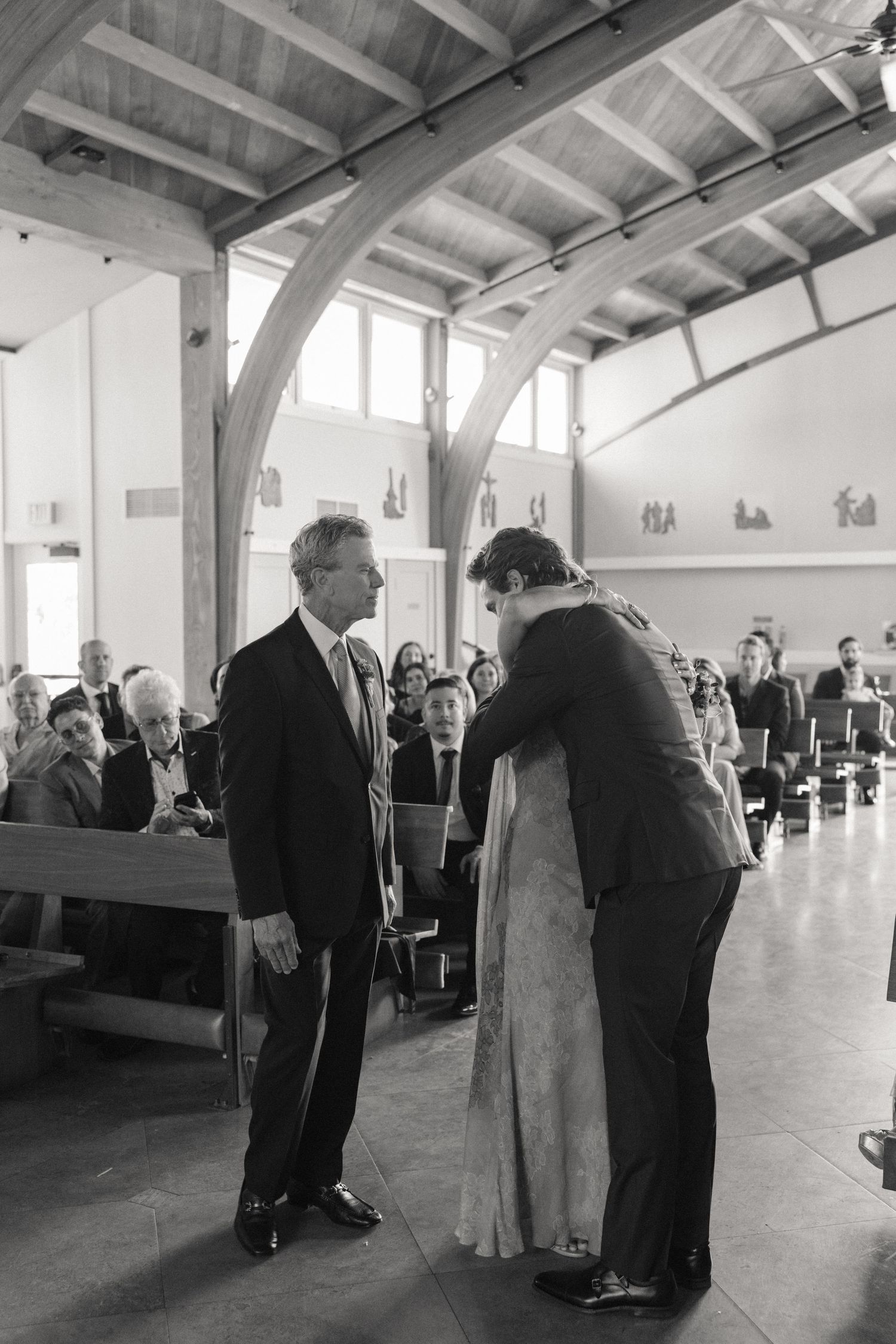 Black and white photo of groom bowing to father during wedding ceremony in modern church.