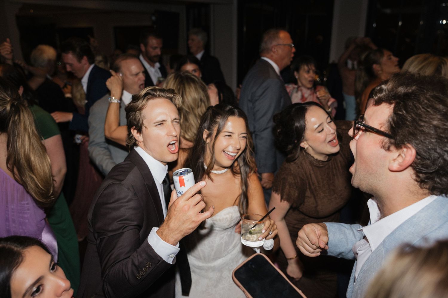 Bride and groom dancing and laughing with guests at a lively indoor wedding reception.