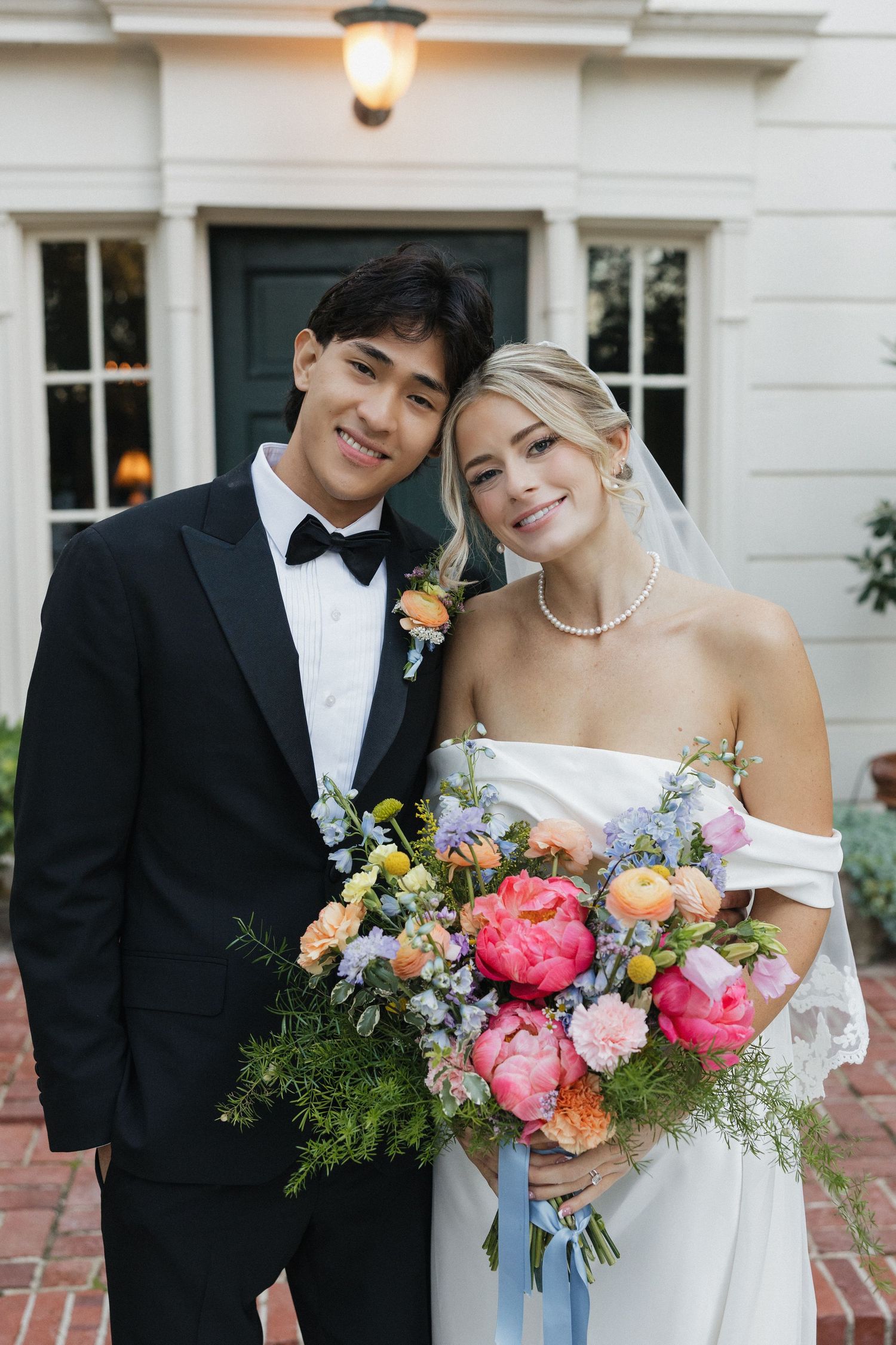 Elegant wedding couple poses with vibrant pink and coral bouquet against white building.