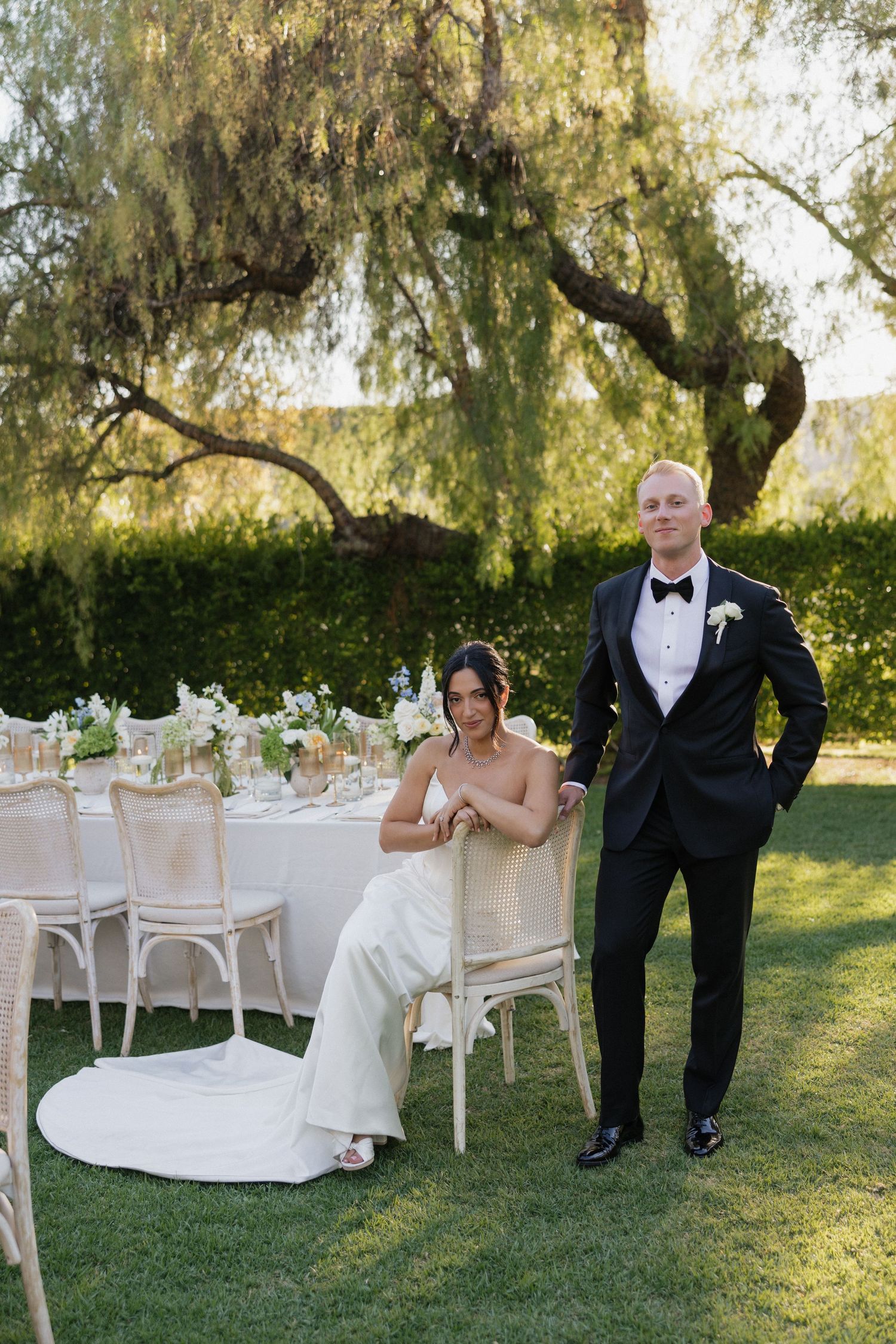 Bride in white gown sits beside groom in black tuxedo at elegant outdoor wedding reception table under willow tree.