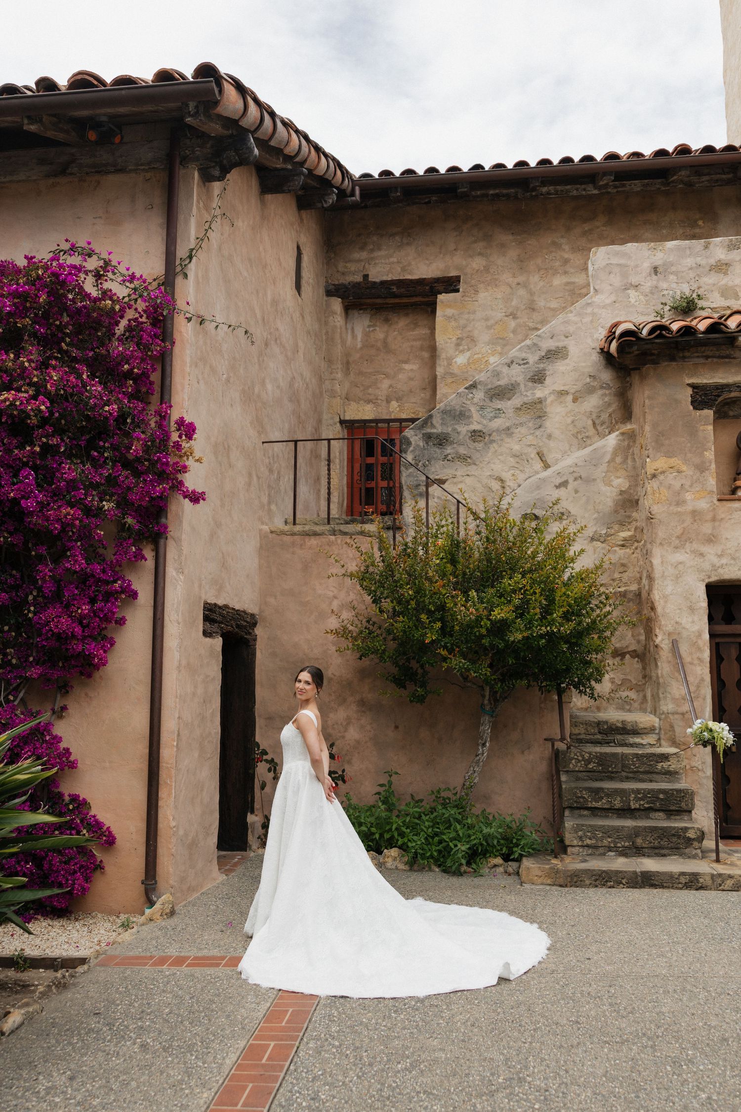 Bride in elegant white gown with long train stands in courtyard of rustic Spanish-style venue with bougainvillea.