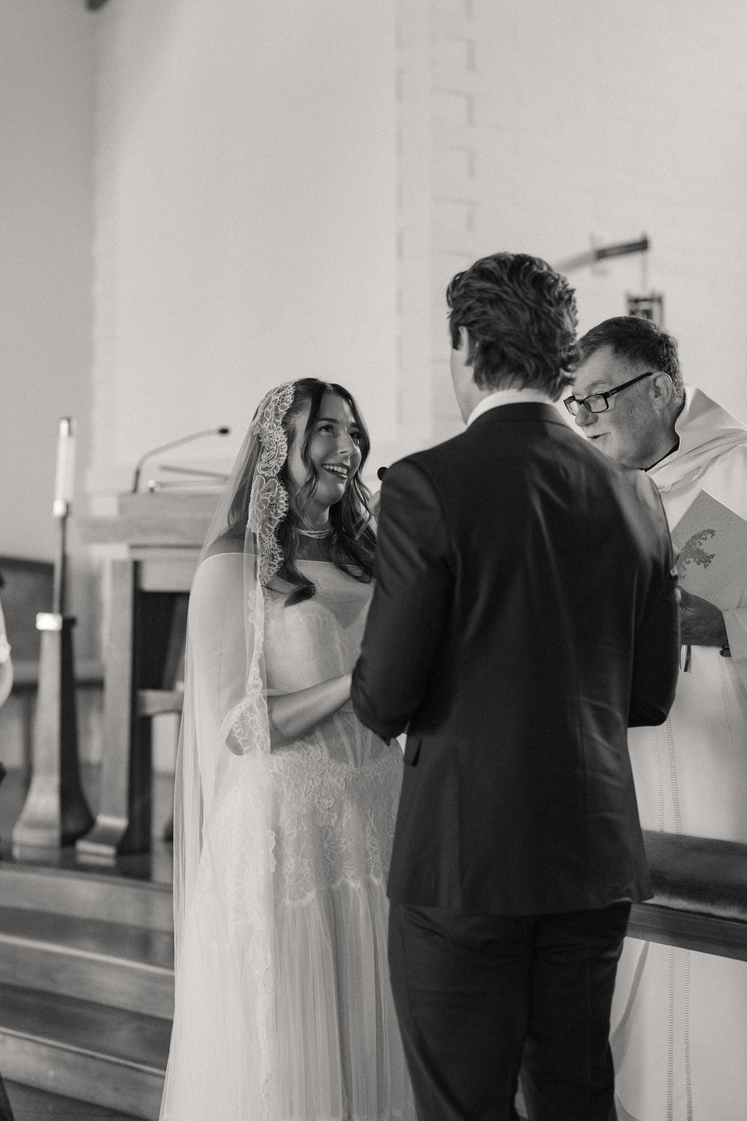 Black and white photo of laughing bride in lace veil holding hands with groom during ceremony.