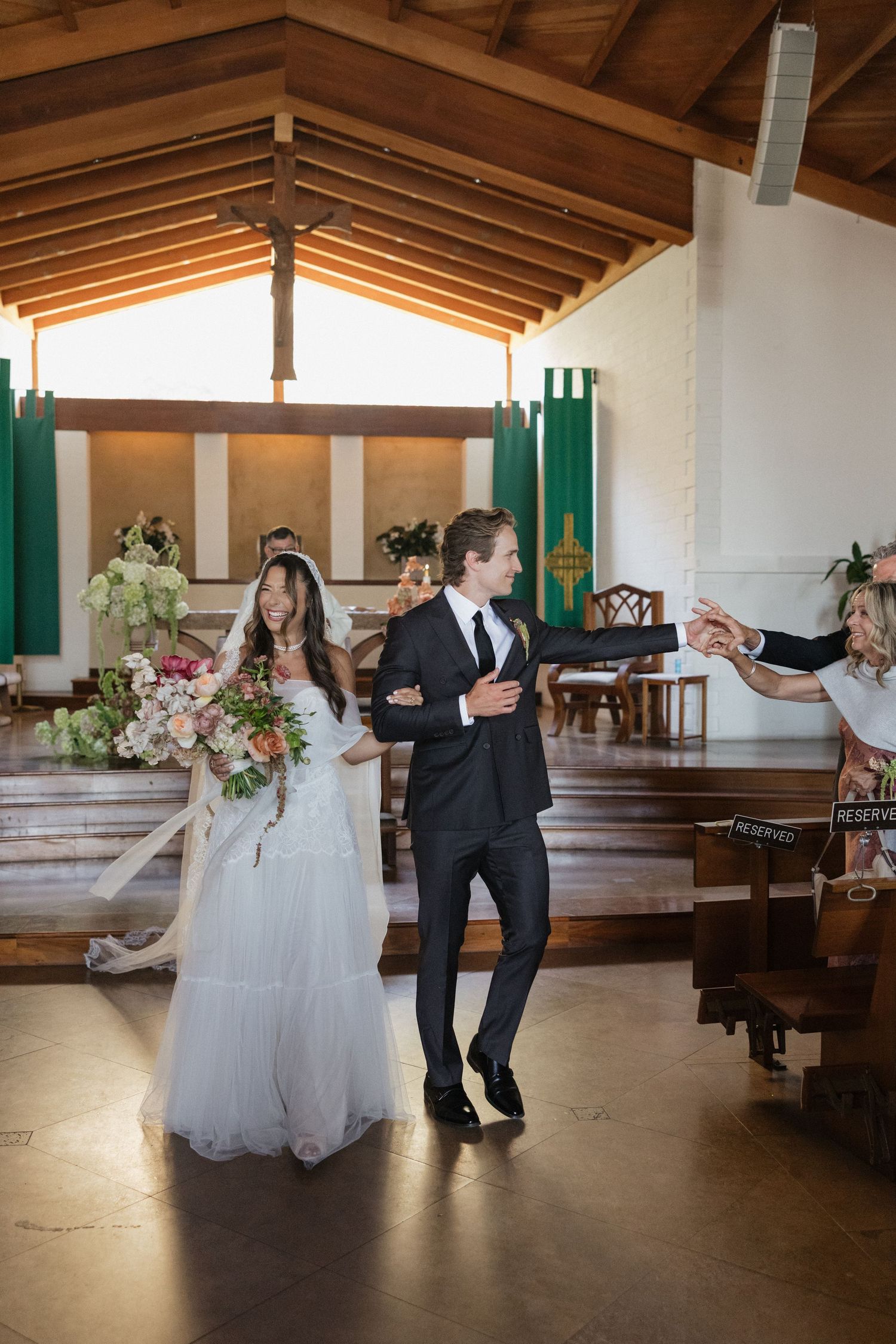 Joyful newlyweds walk down church aisle after ceremony, bride holding colorful bouquet with long veil.