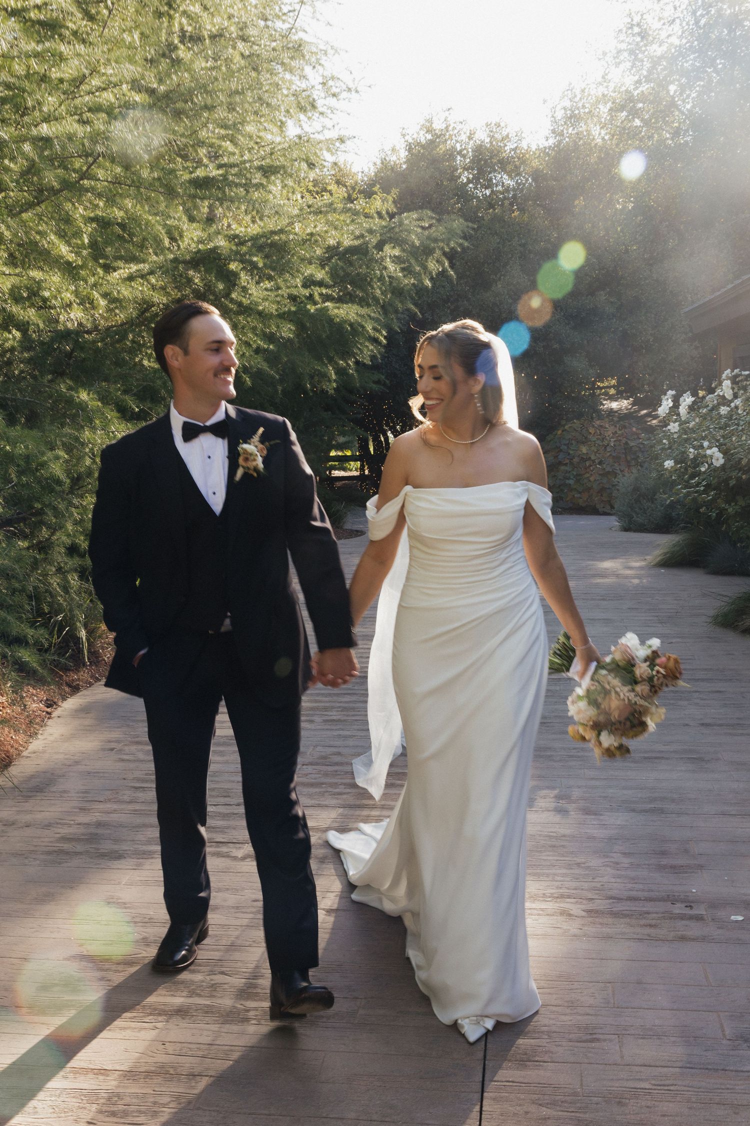 Newlyweds walk together on stone path surrounded by trees with sunlight streaming through.