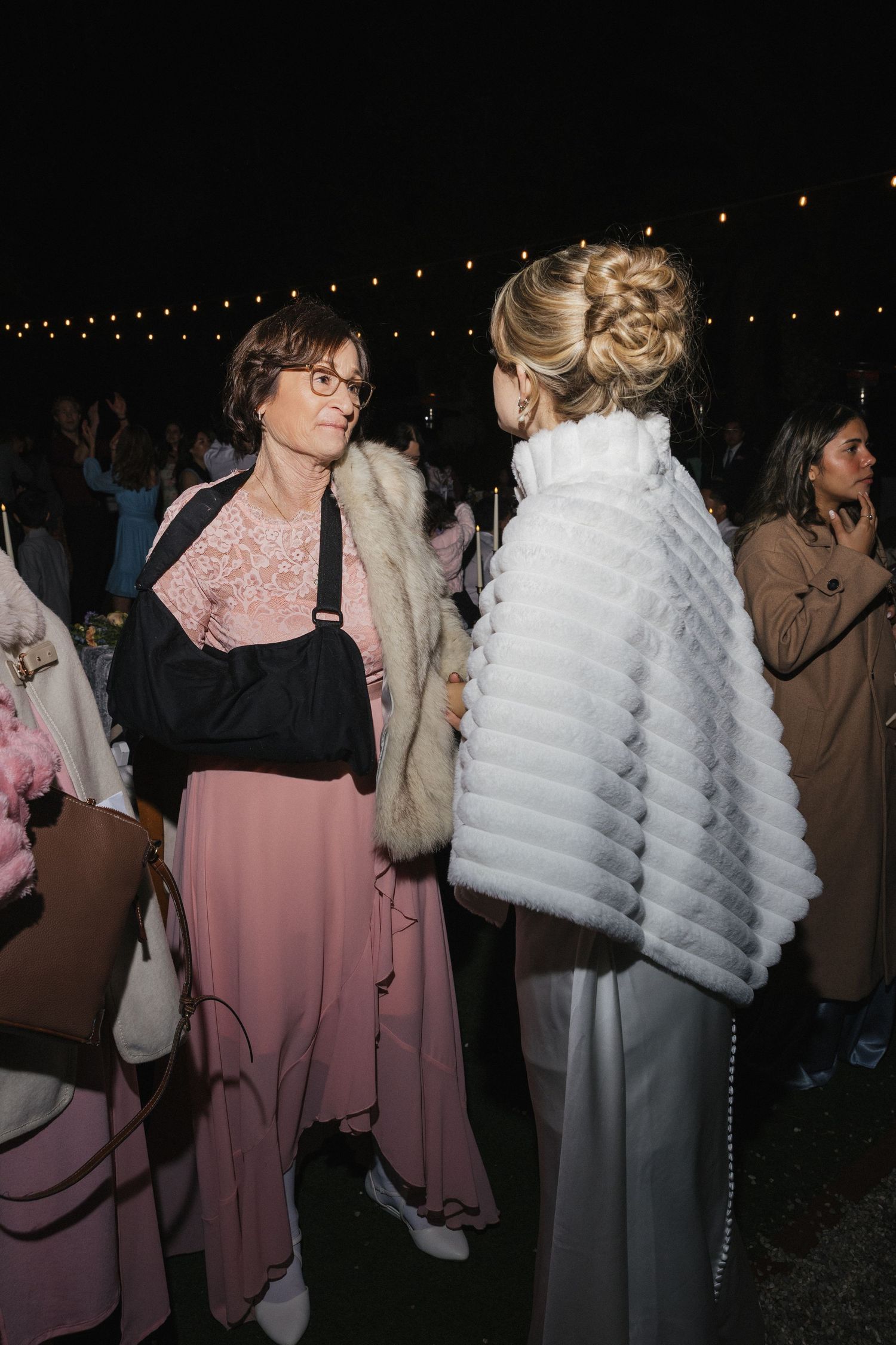 Wedding guests in pink dresses and white fur shawls mingle at an evening reception.