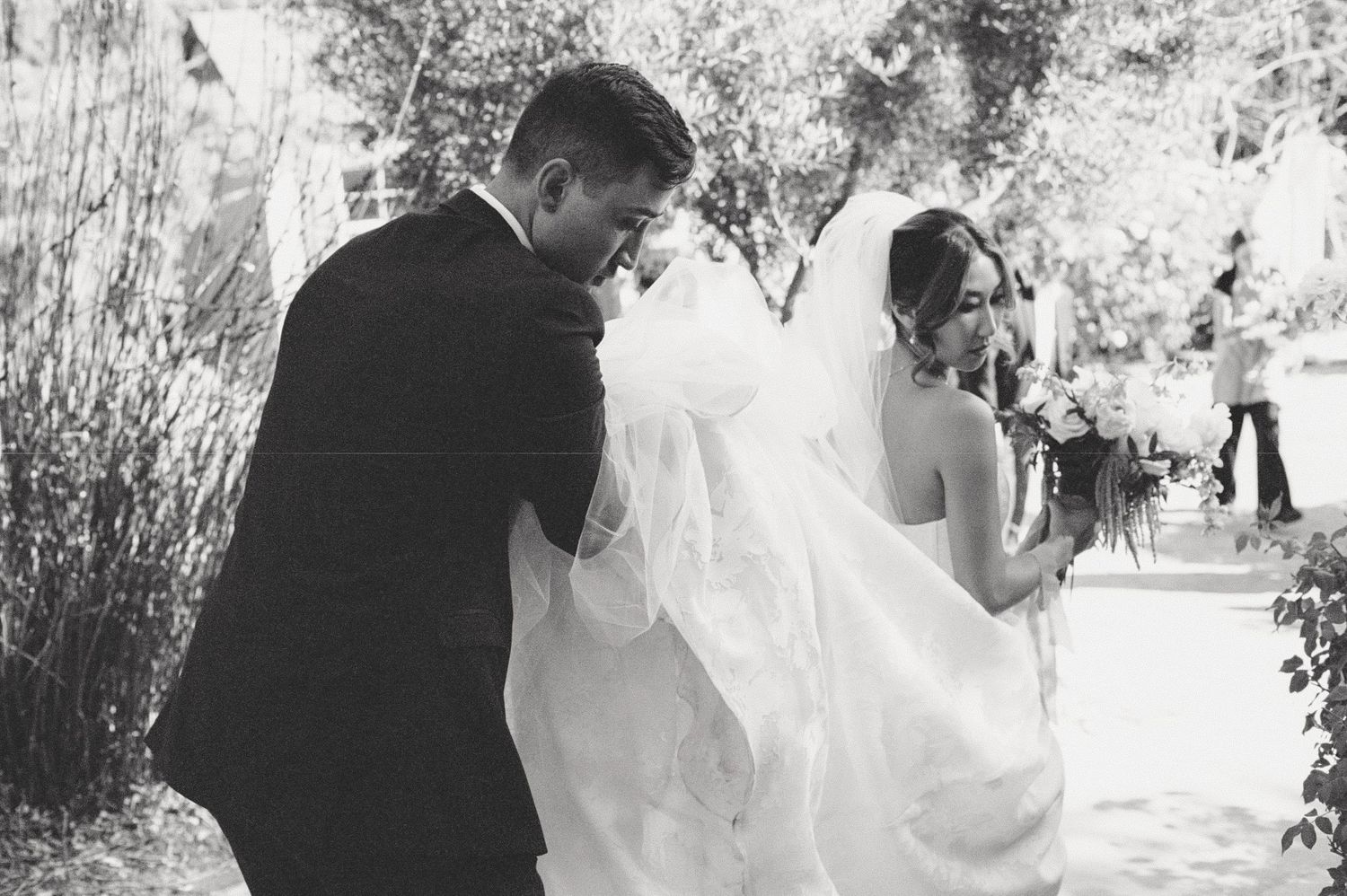 Elegant black and white wedding photo of a couple walking together with flowing veil in natural setting.