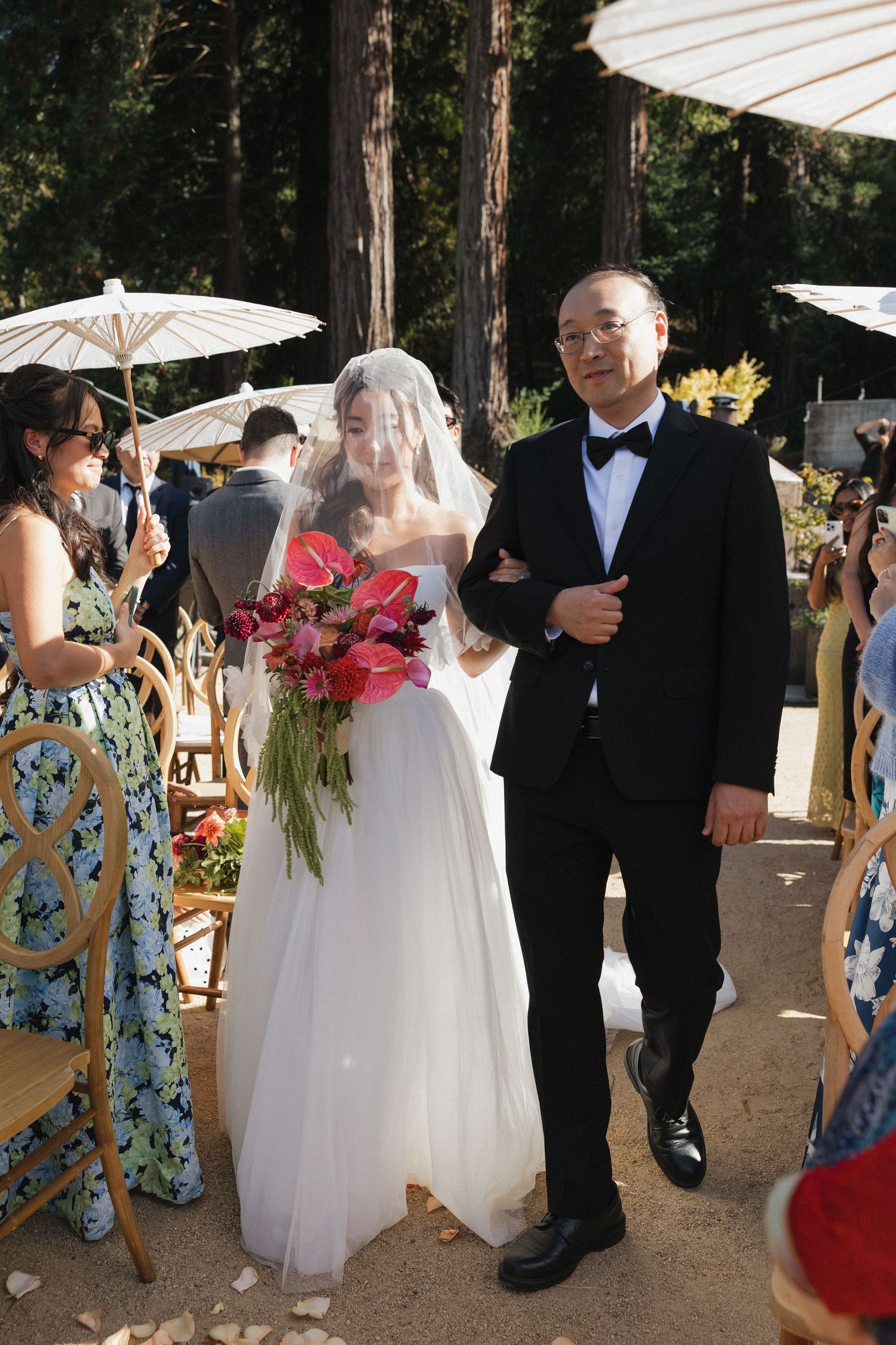 Wedding guests gather under white umbrellas during an outdoor ceremony among tall trees.